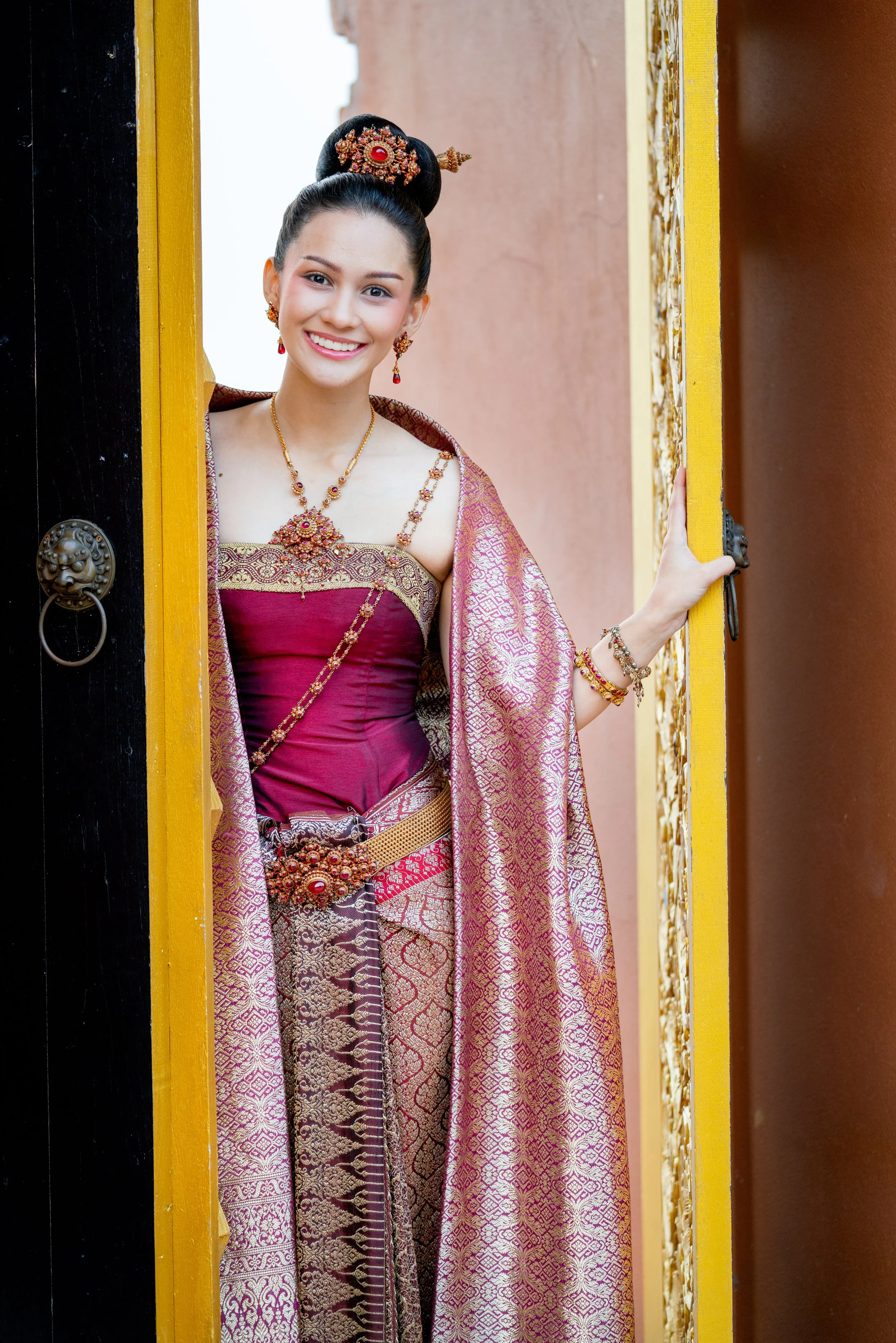 Young woman smiling and wearing traditional Thai attire, standing at a doorway.