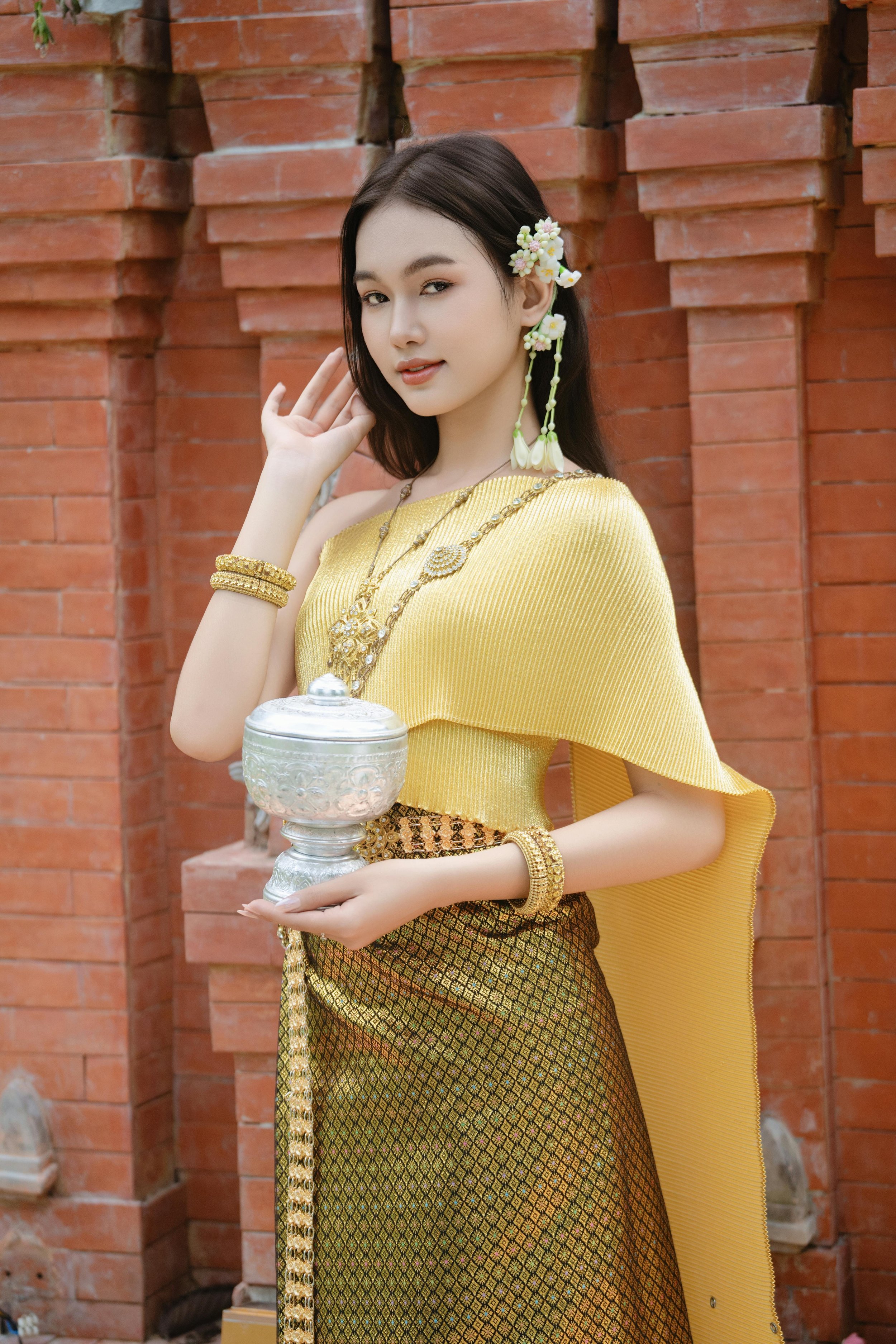 A woman dressed in traditional Thai attire, holding a silver container, standing in front of a brick wall, with jewelry and floral accessories.