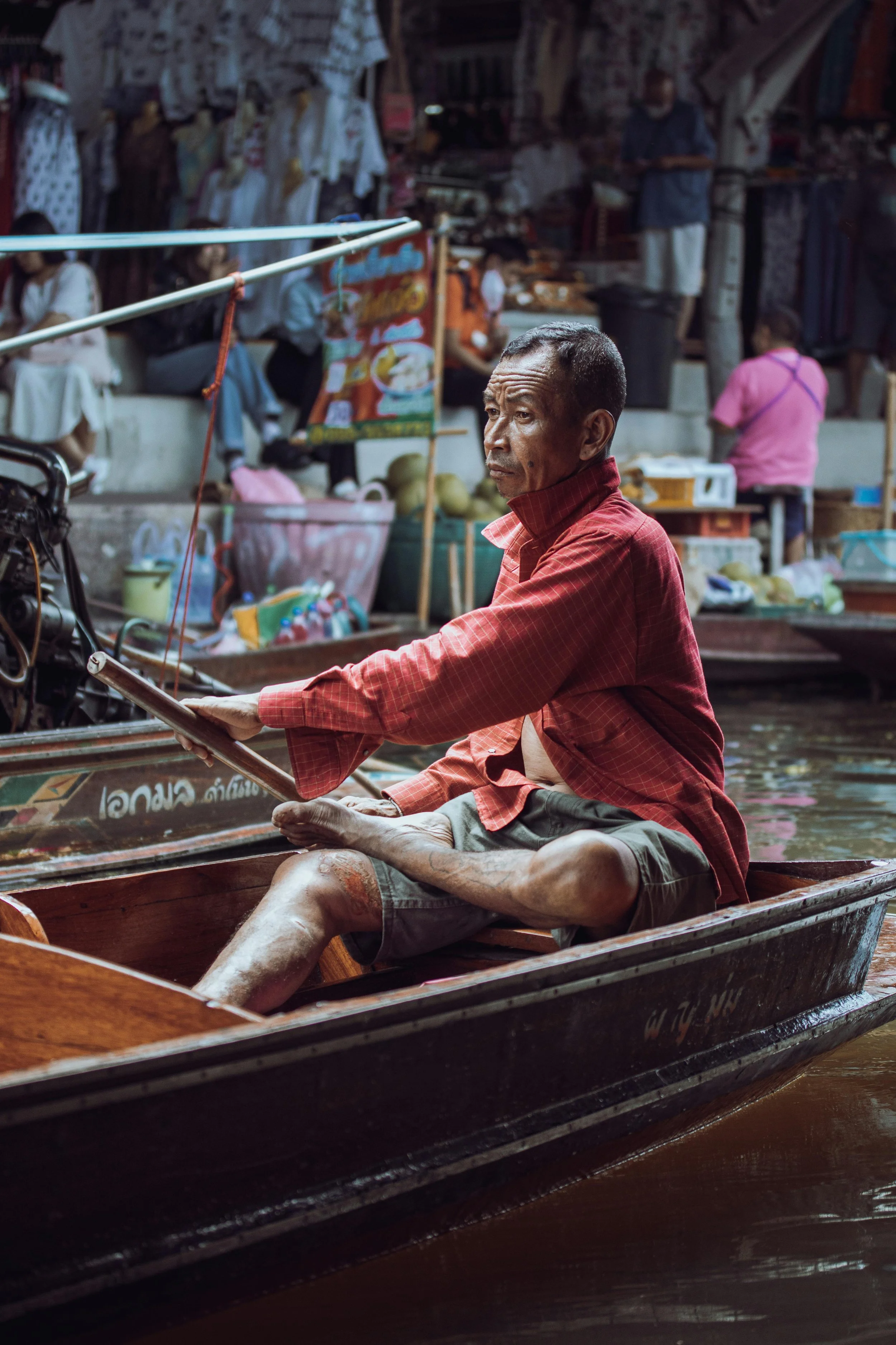 A man sitting in a wooden boat on a canal market, holding a tablet, with market stalls and people in the background.
