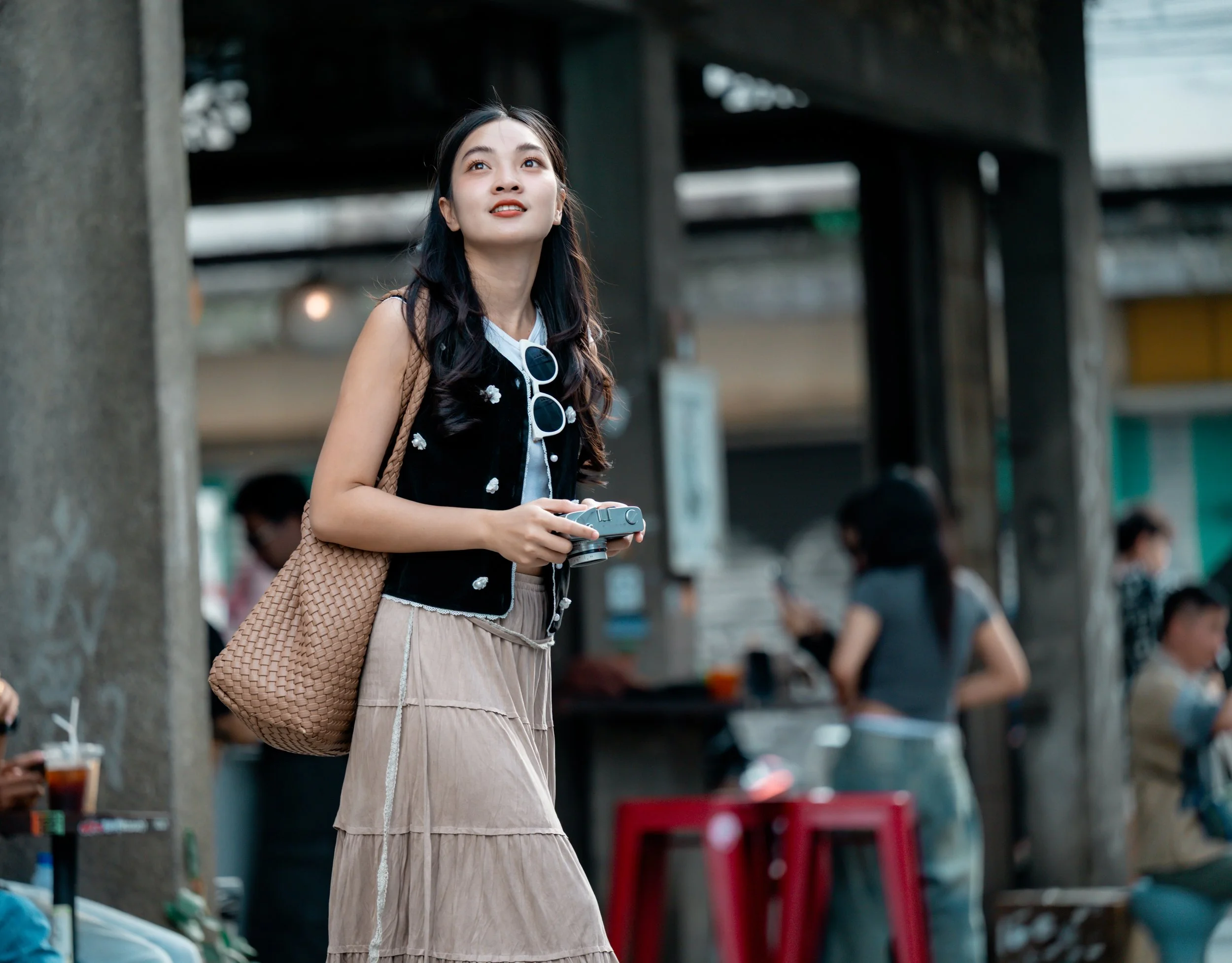 A young woman with long dark hair, carrying a woven tan bag and holding a vintage camera, walking outdoors in an urban setting on a cloudy day.