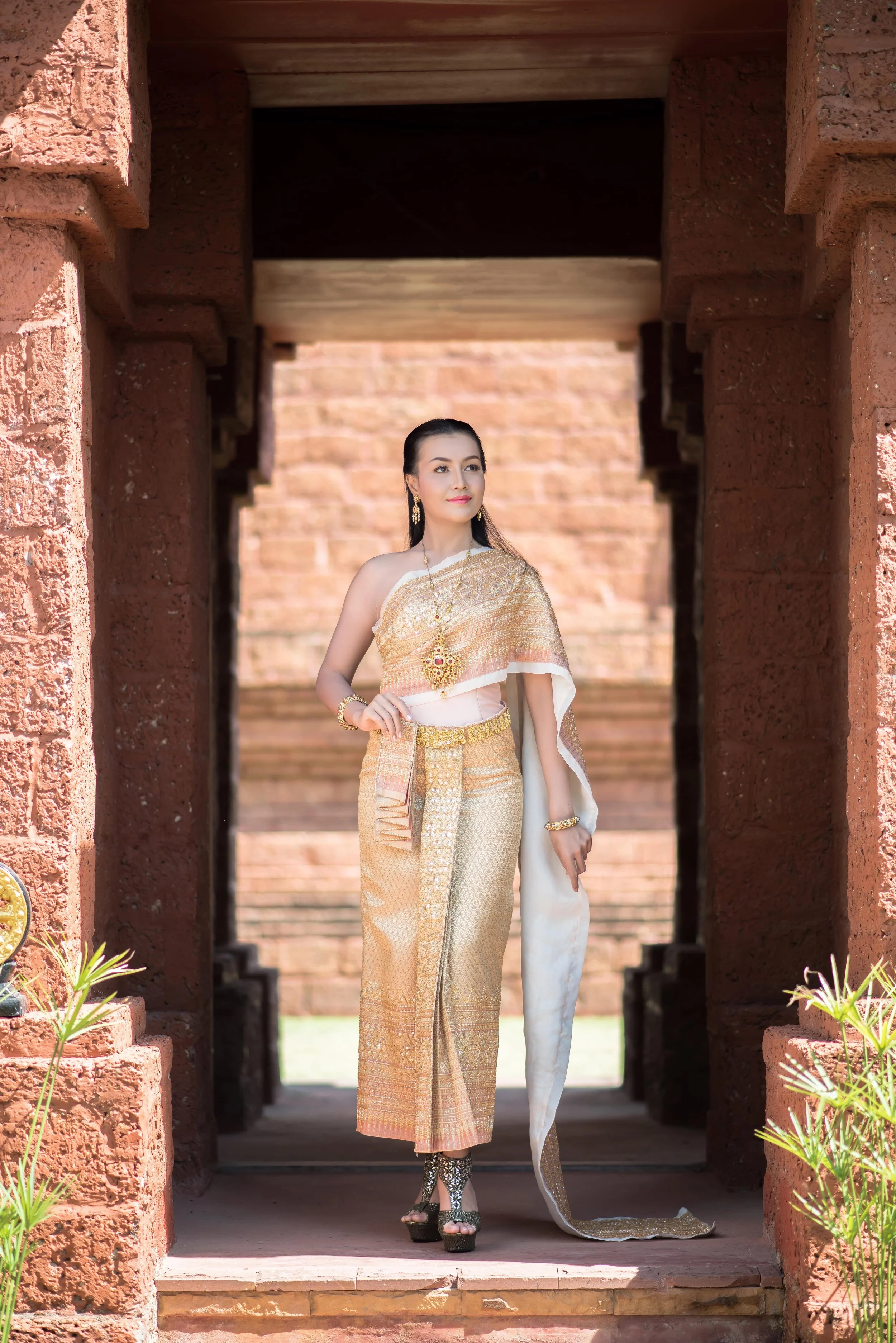 A woman dressed in traditional Thai attire standing in a brick archway with ancient ruins in the background.