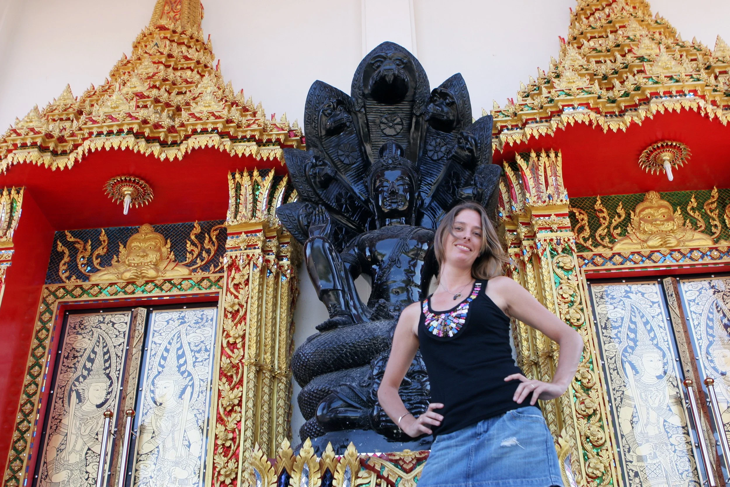 A woman posing in front of a large, ornate statue of a multi-headed serpent with lion-like faces, set against a decorated, traditional Thai style temple with gold accents, intricate patterns, and mythical creature motifs.