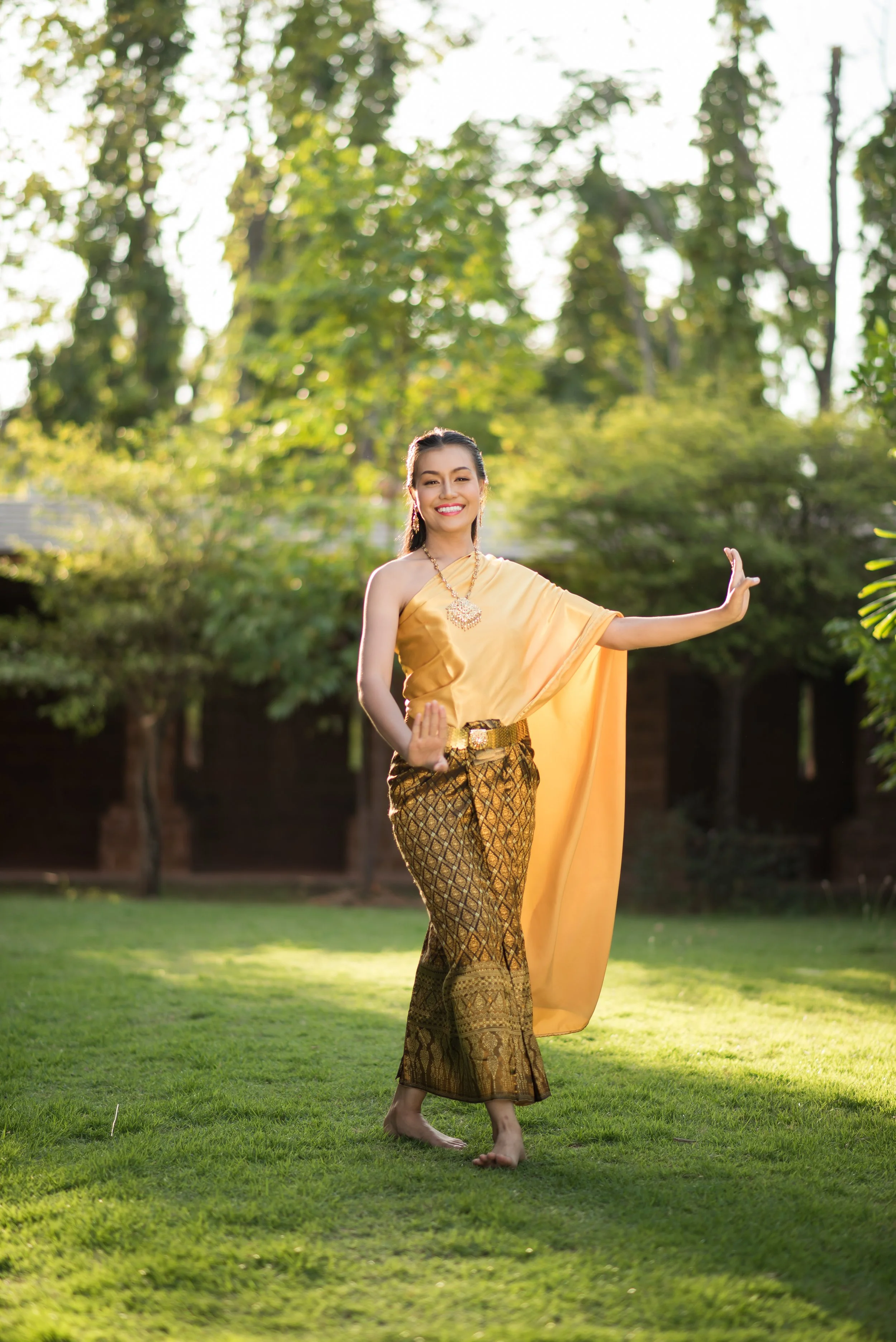 A woman dressed in traditional Thai attire, smiling, posing with one arm extended and the other bent, outdoors on green grass with trees in the background.
