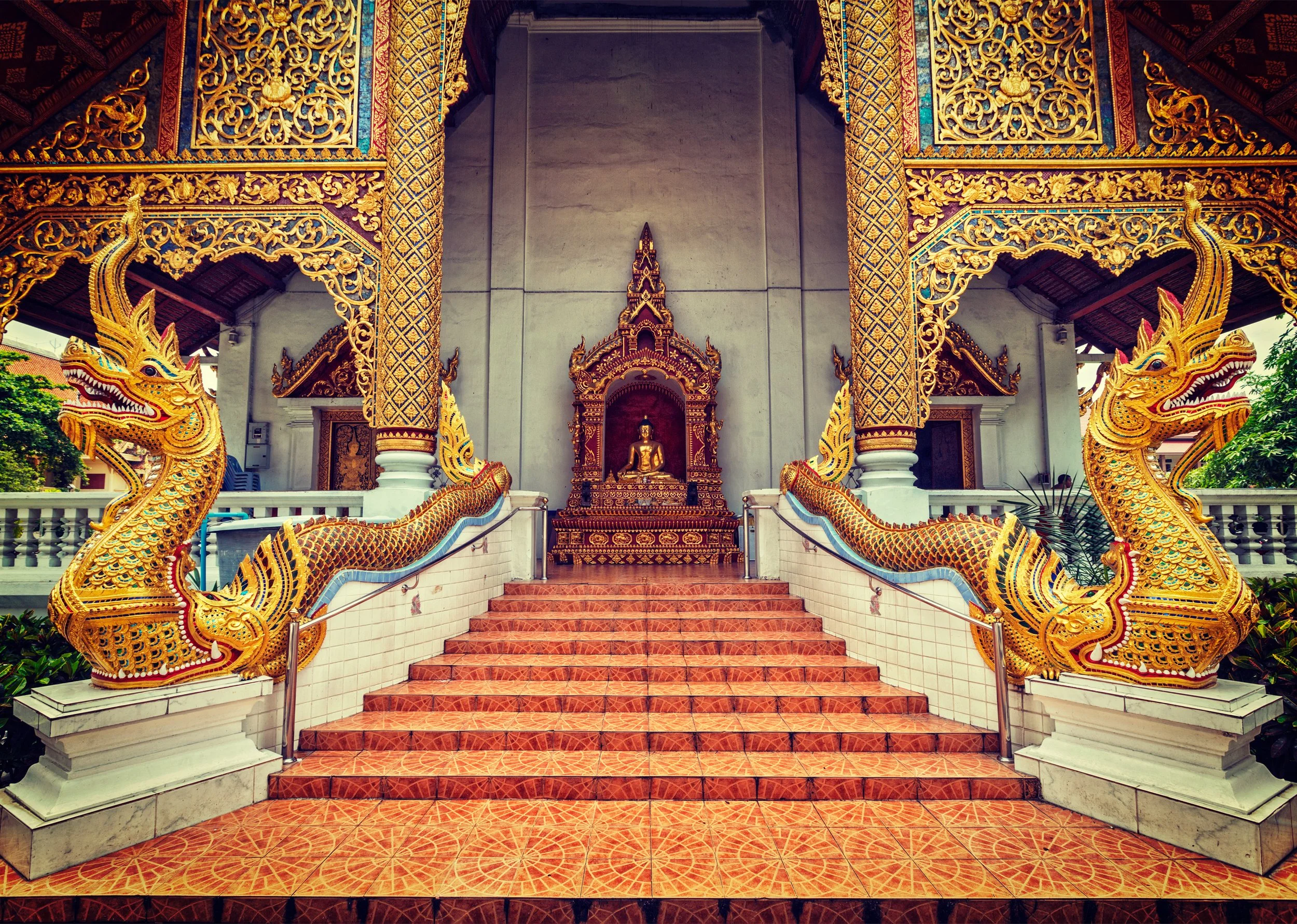 A temple entrance with a statue of Buddha at the top of a red staircase, flanked by two golden dragon-shaped sculptures.