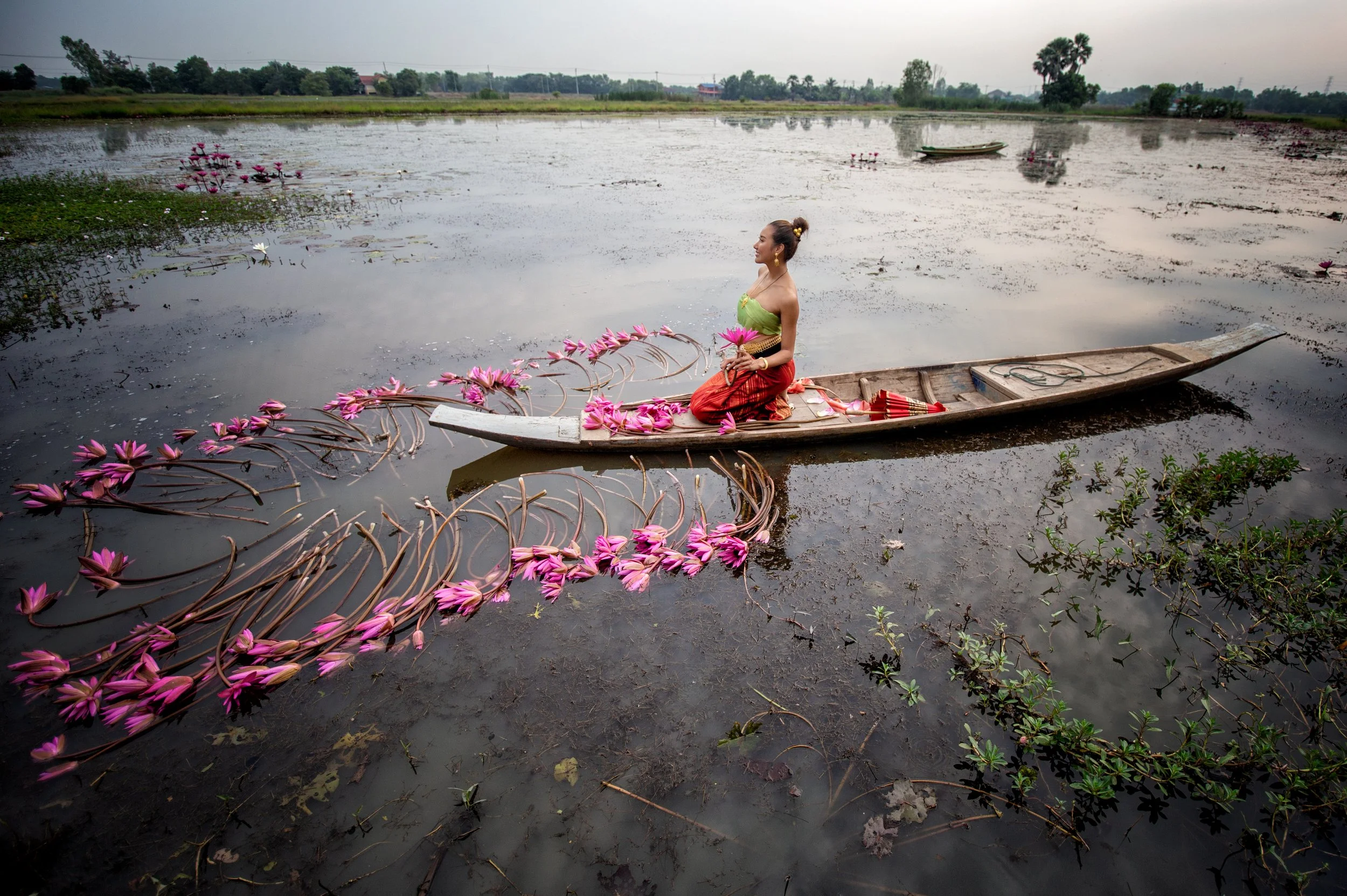 A woman in traditional clothing sitting in a boat on a water body surrounded by pink water lilies.