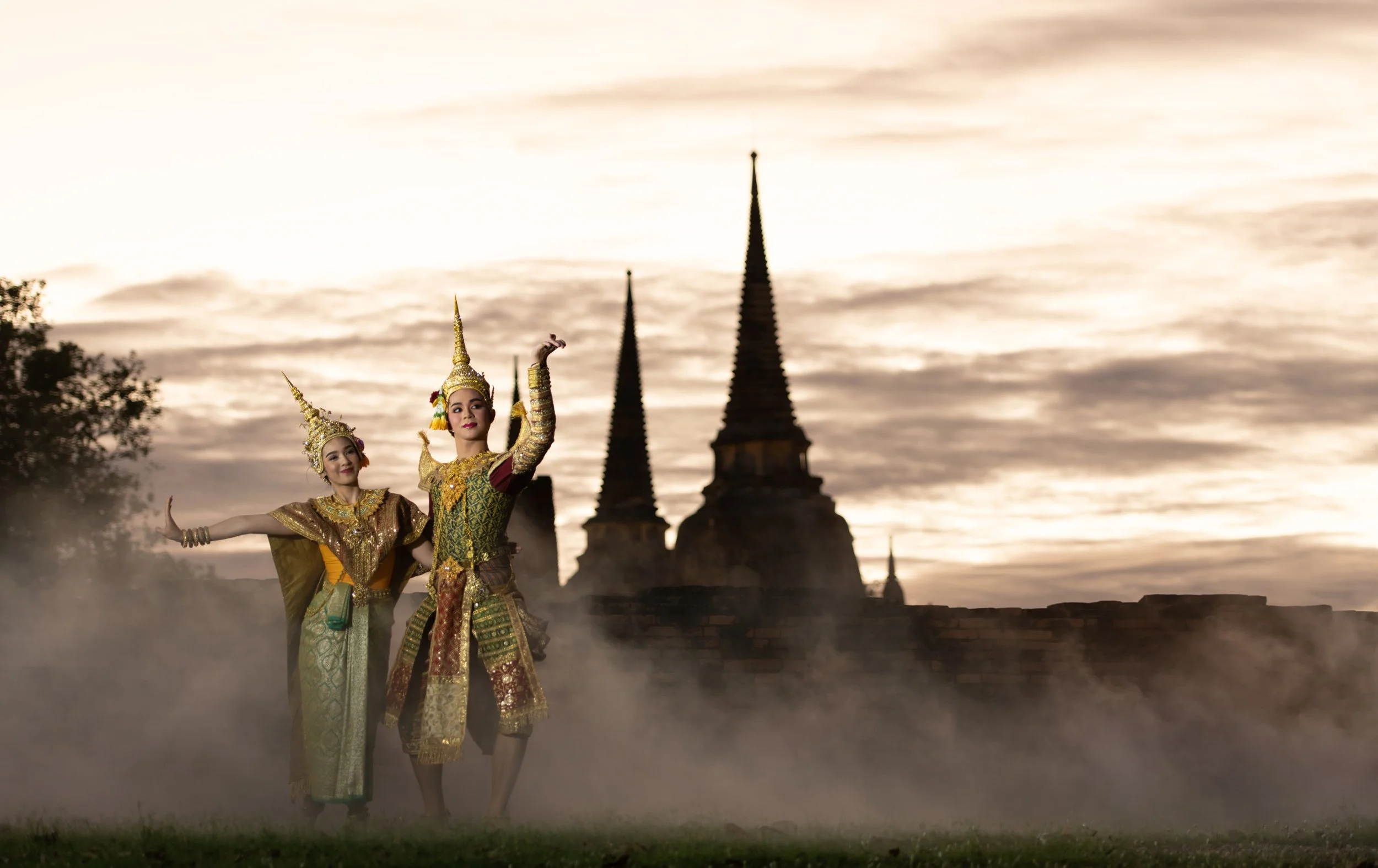 Two women in traditional Thai dance costumes performing in front of ancient temple ruins during sunset.