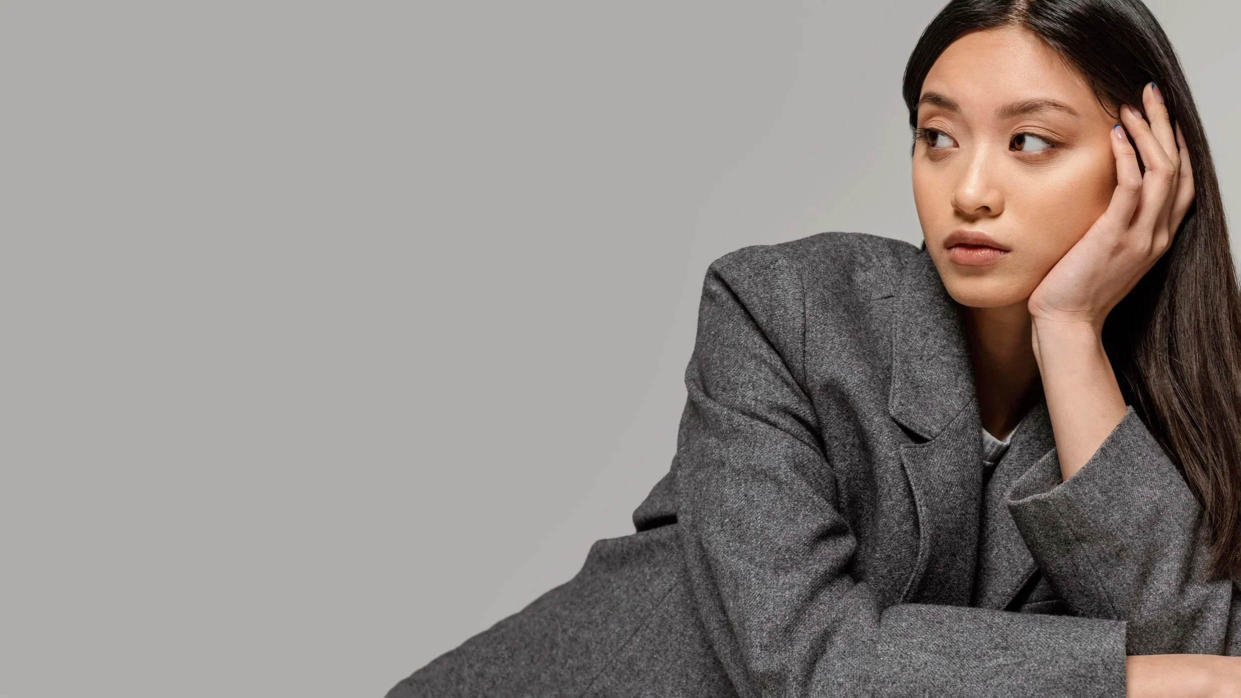 A young woman with long dark hair in professional gray blazer looking thoughtfully to the side, resting her head on her hand, against a plain gray background.