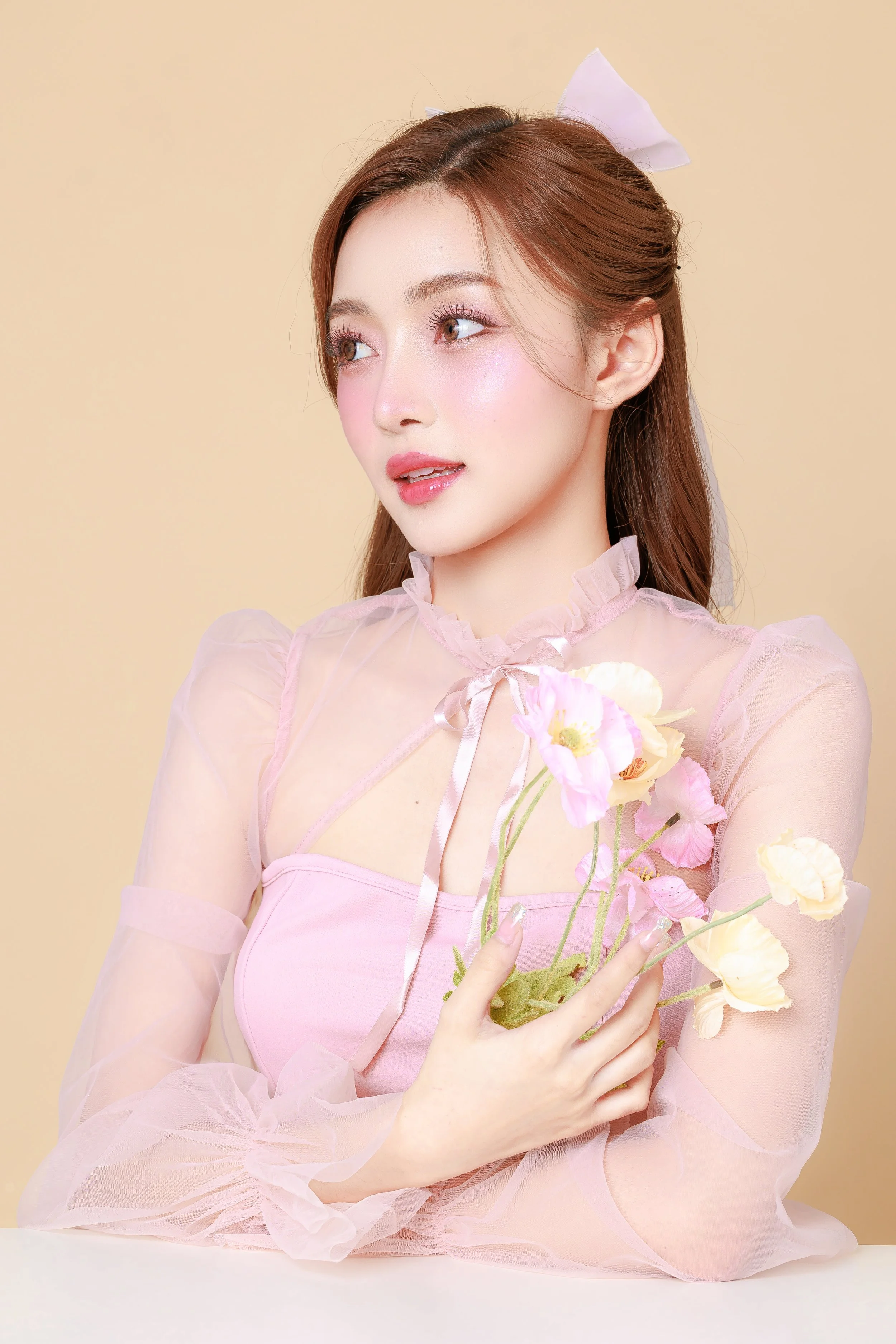 A woman with long brown hair and light makeup, wearing a pink sheer blouse with ruffled details and a bow, holding a small bouquet of soft pink and yellow poppies, standing against a plain beige background.