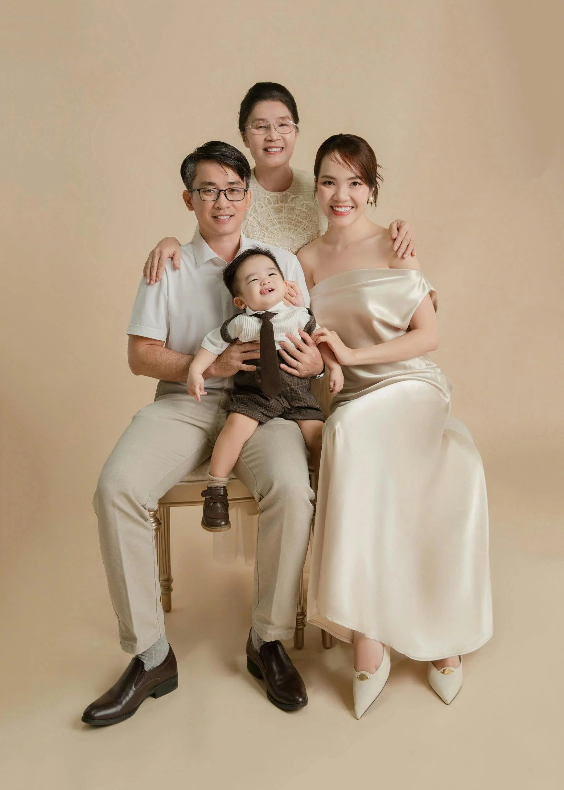 A multi-generational Asian family portrait, including an elderly woman, a young woman, a young man, and a young boy, dressed in elegant and casual clothing against a beige background, smiling and close together.
