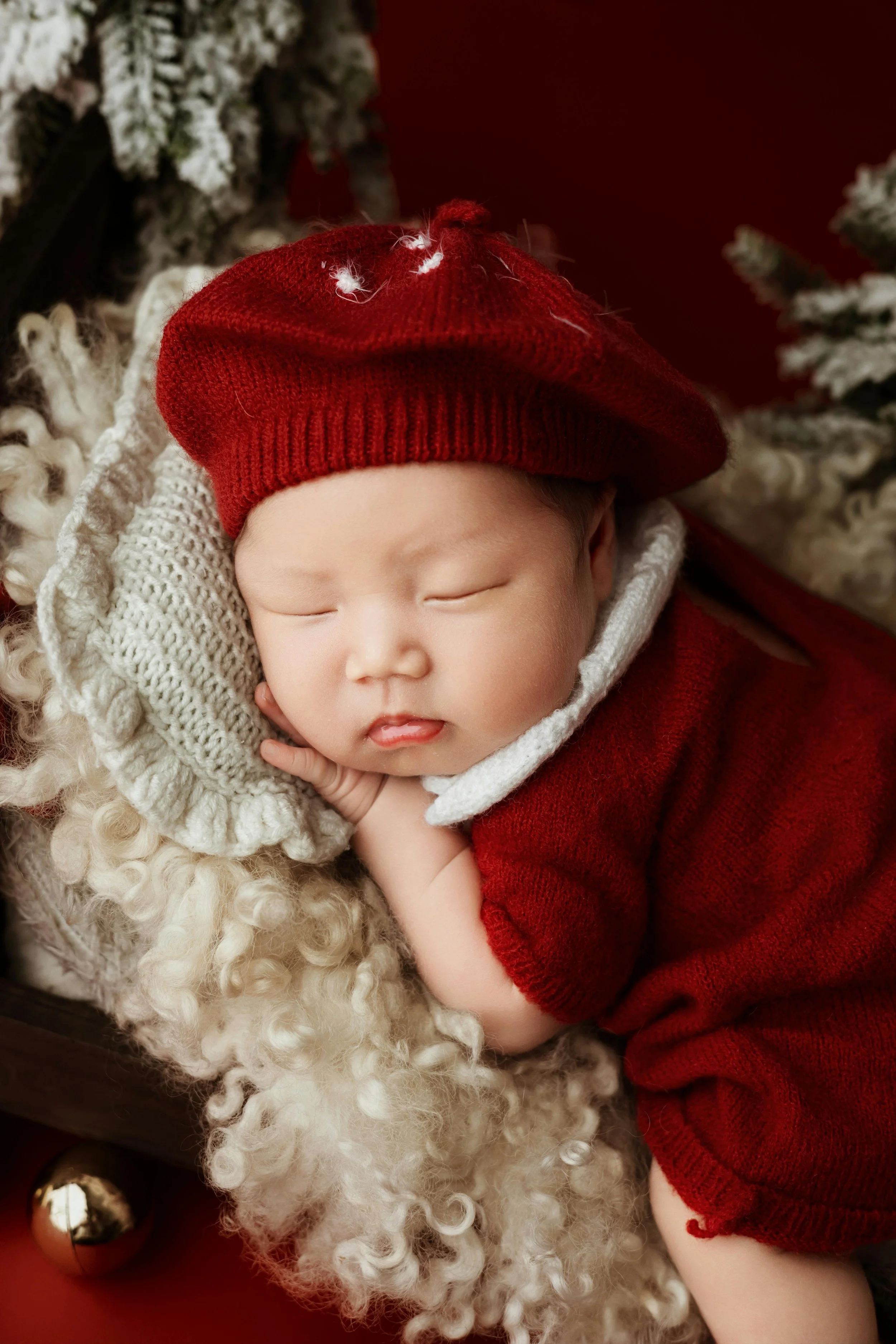 A sleeping baby dressed in a red knit hat and matching red outfit, lying on a cream-colored, curly faux fur blanket.