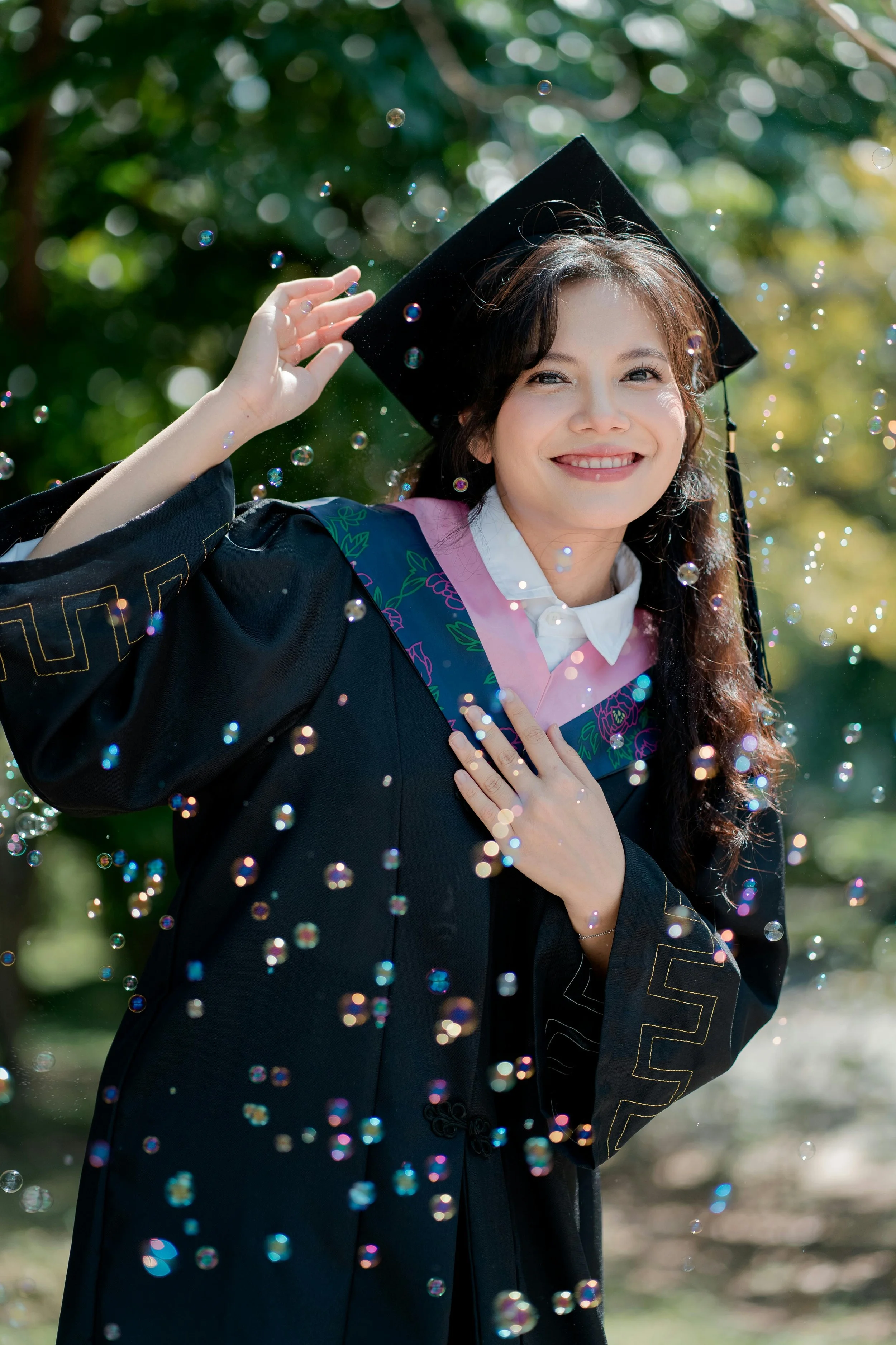 A young woman in a graduation cap and gown smiling outdoors surrounded by floating bubbles.