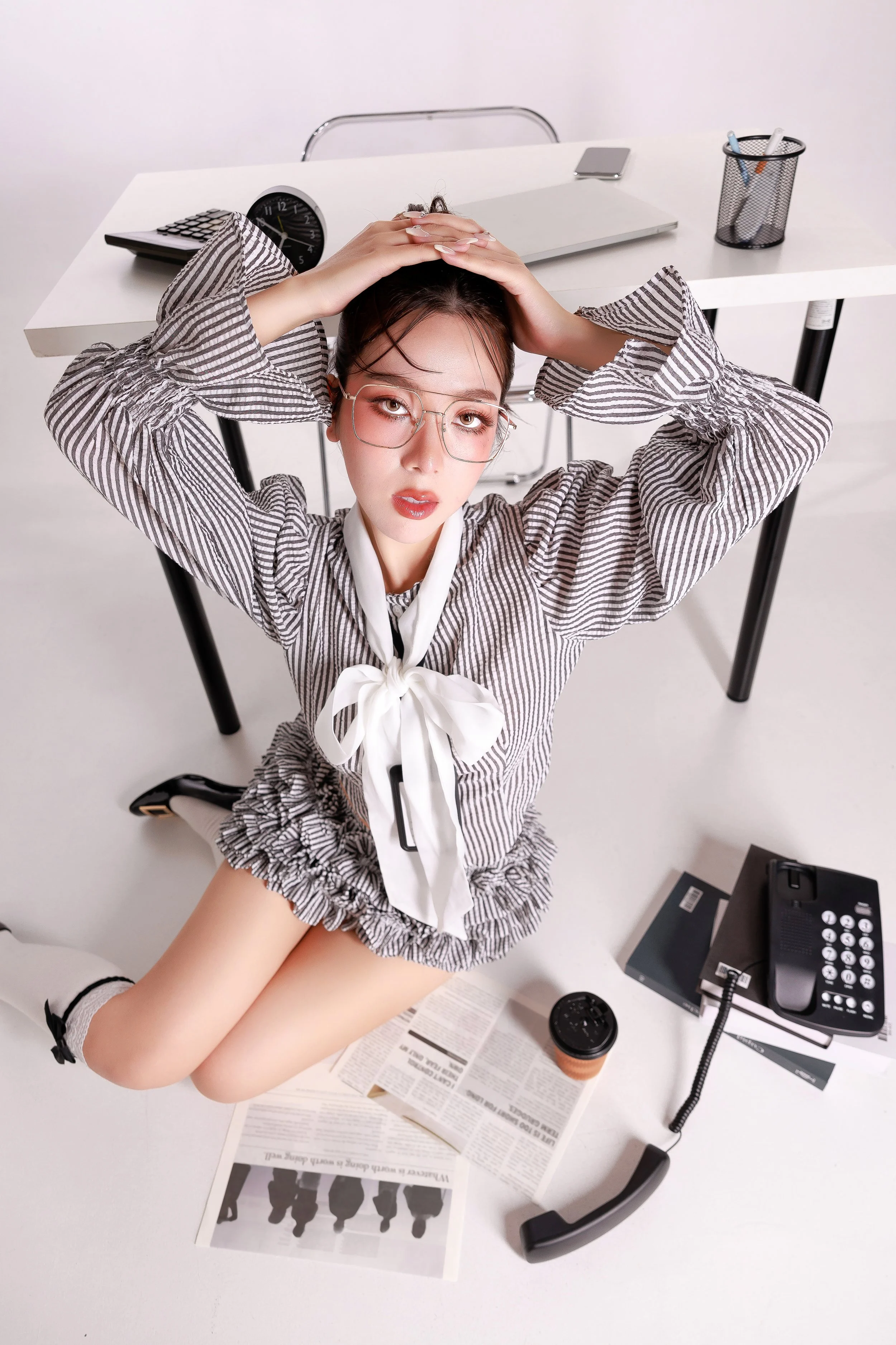 Young woman in office attire kneeling on a desk, looking up at camera with hands on her head, surrounded by office items including a telephone, papers, a coffee cup, and a laptop.