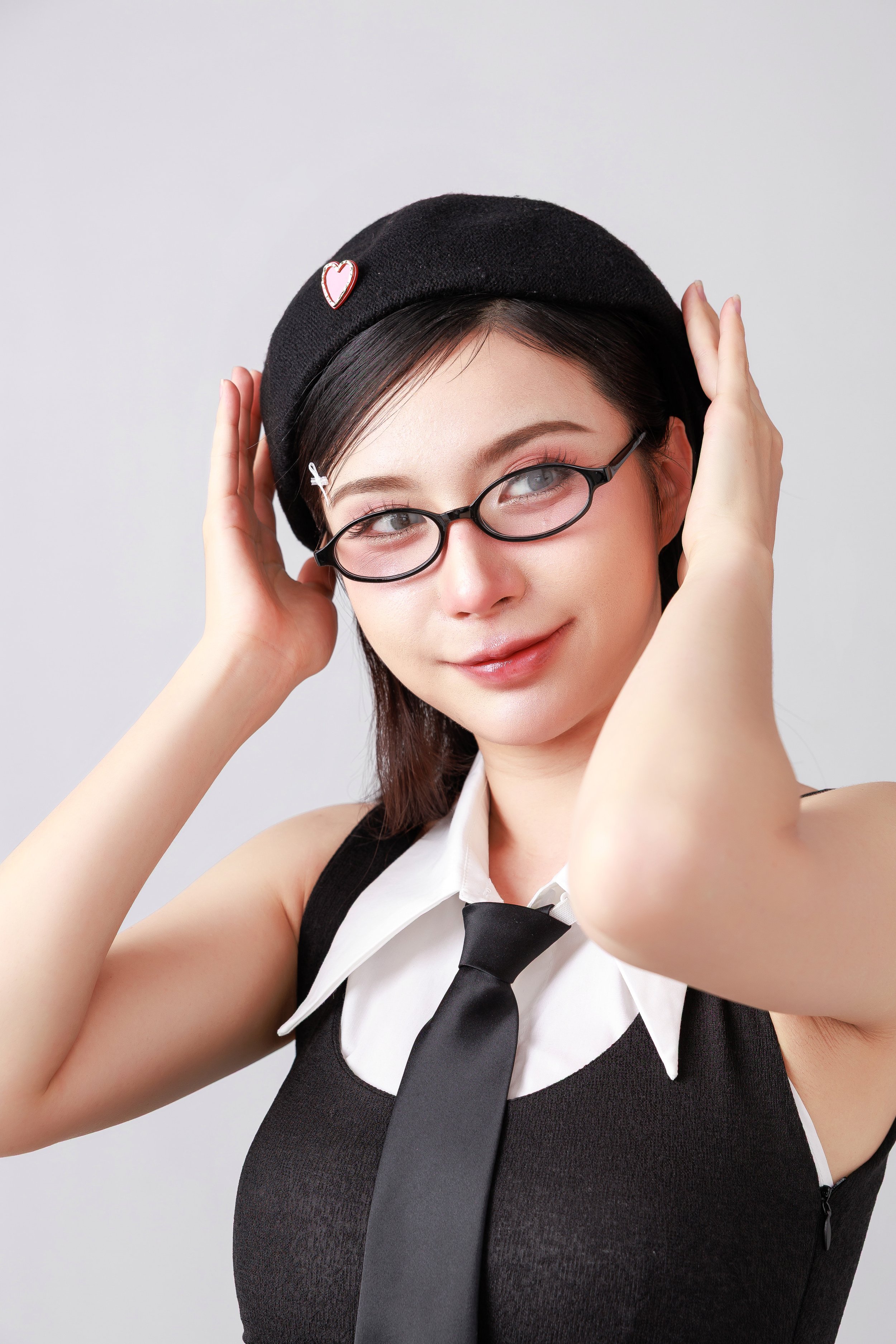 A young woman with glasses, wearing a black beret with a heart pin, a white collared shirt, a black tie, and a black sleeveless top, smiling and adjusting her beret.