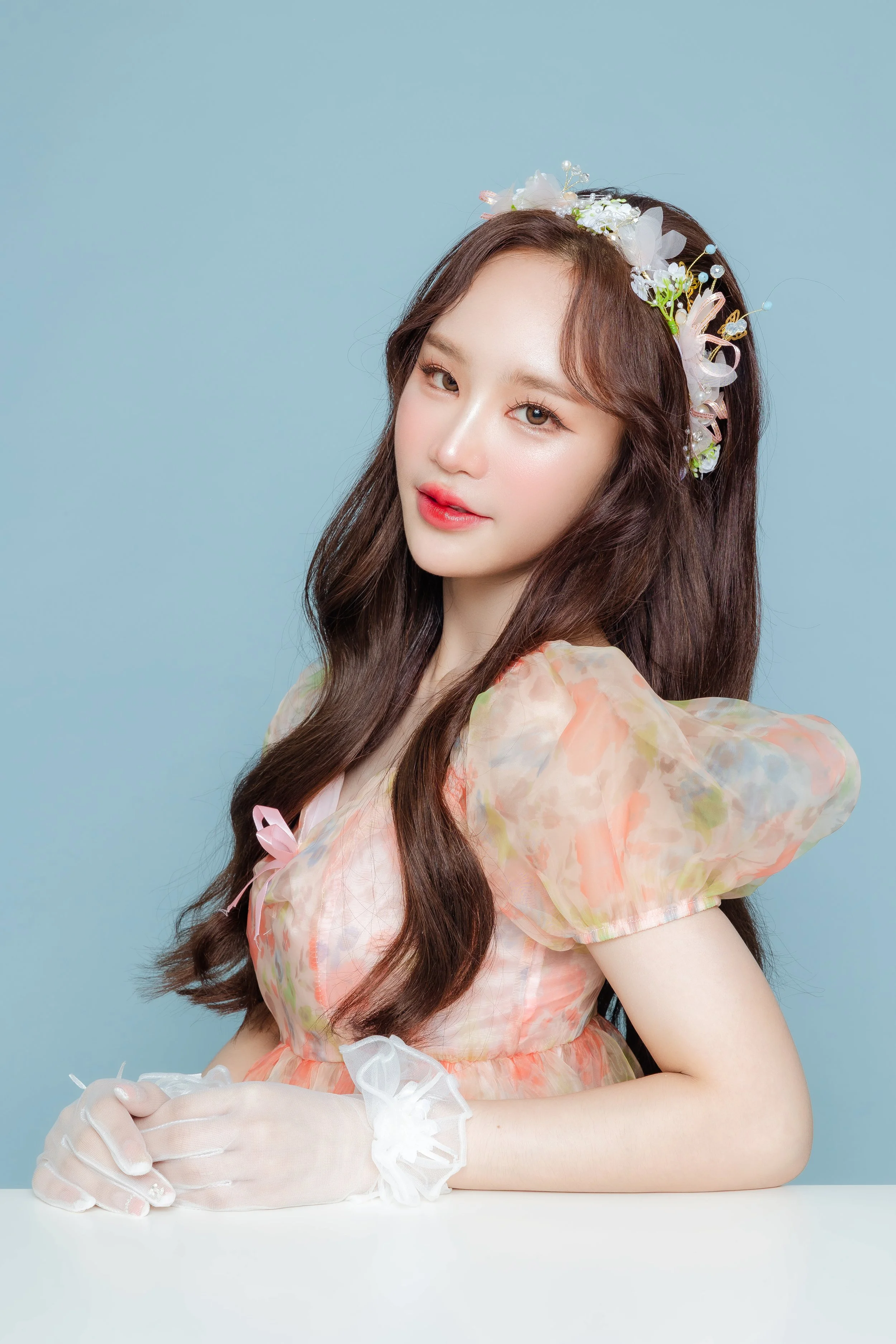 A young woman with long brown hair wearing a floral dress and a decorative headband with flowers and beads, posed against a light blue background.