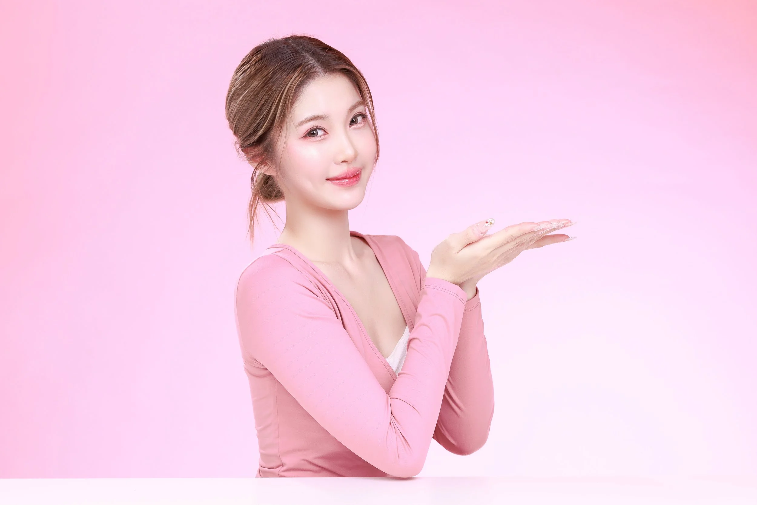 A young woman with light brown hair in a loose bun, wearing a pink long-sleeve shirt, smiling and holding her hands together in a cupped position against a pink gradient background.