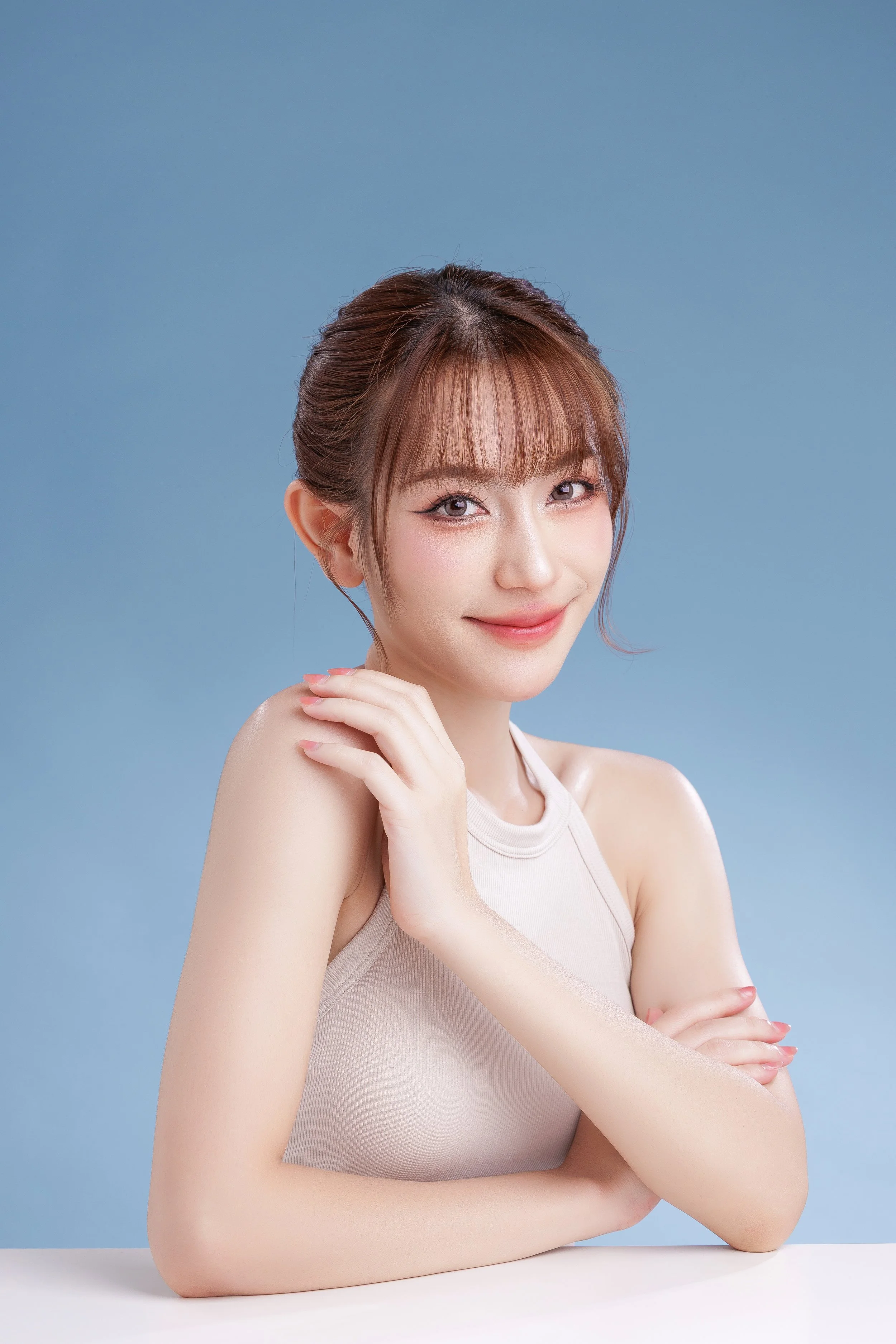 A young woman with light skin and brown hair styled in an updo, smiling softly at the camera, with her arms crossed and resting on a white surface, against a light blue background.