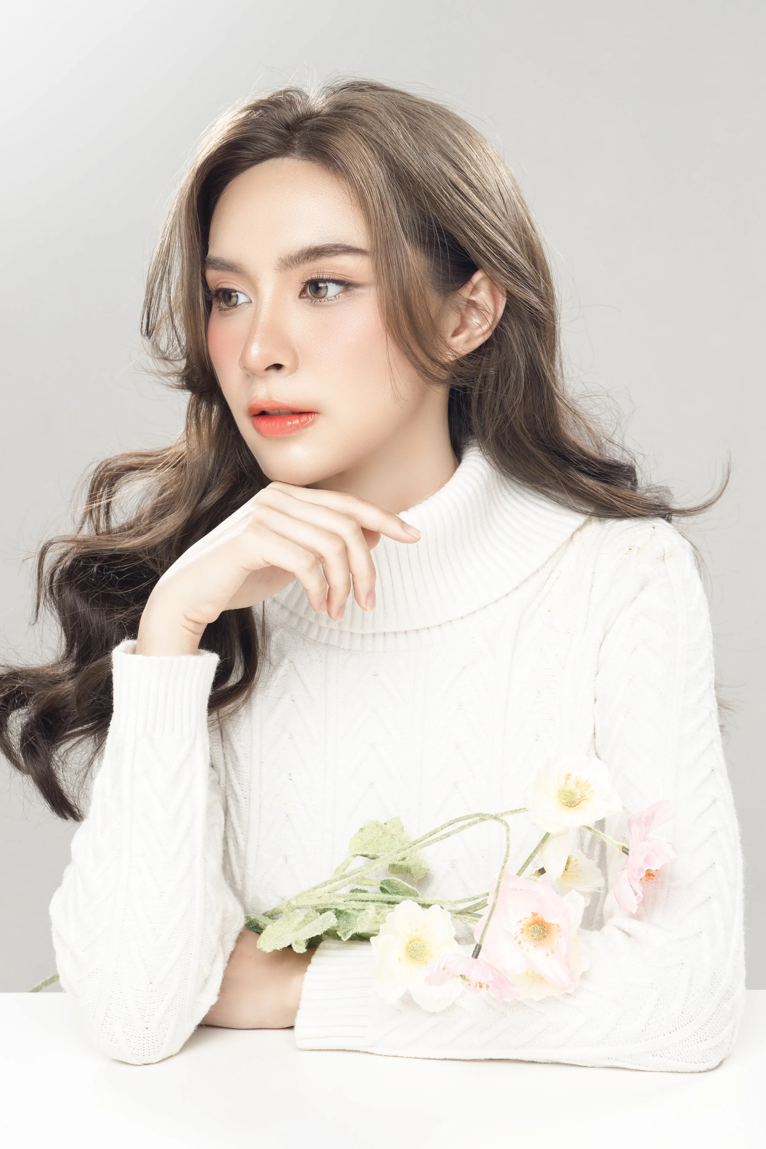 A woman with long wavy brown hair, wearing a white turtleneck sweater, holding pink and white flowers, looking thoughtful against a light gray background.