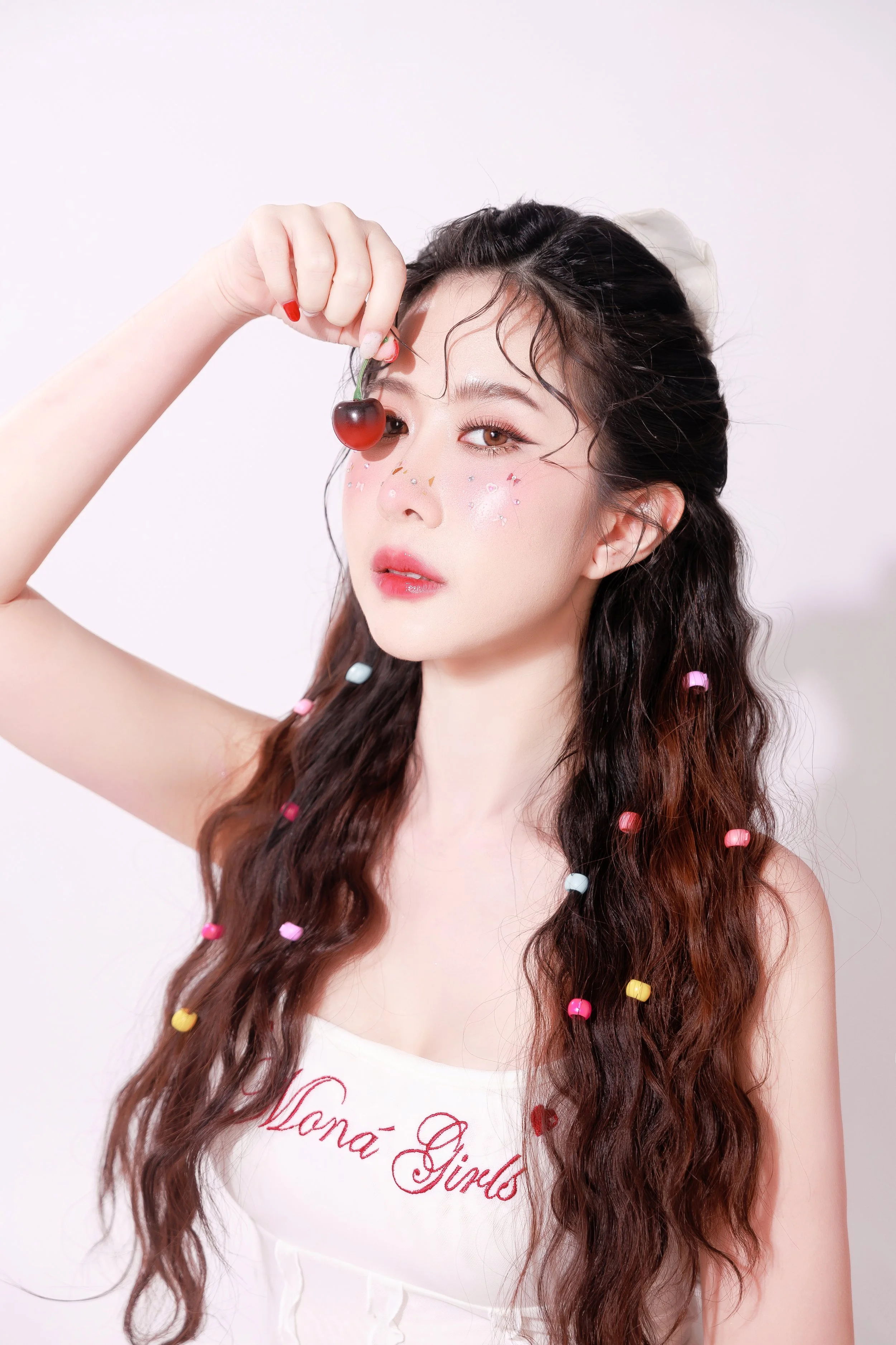 A young woman with long, wavy brown hair decorated with colorful beads, holding a cherry near her eye, with decorative stickers on her face, posing against a plain white background.