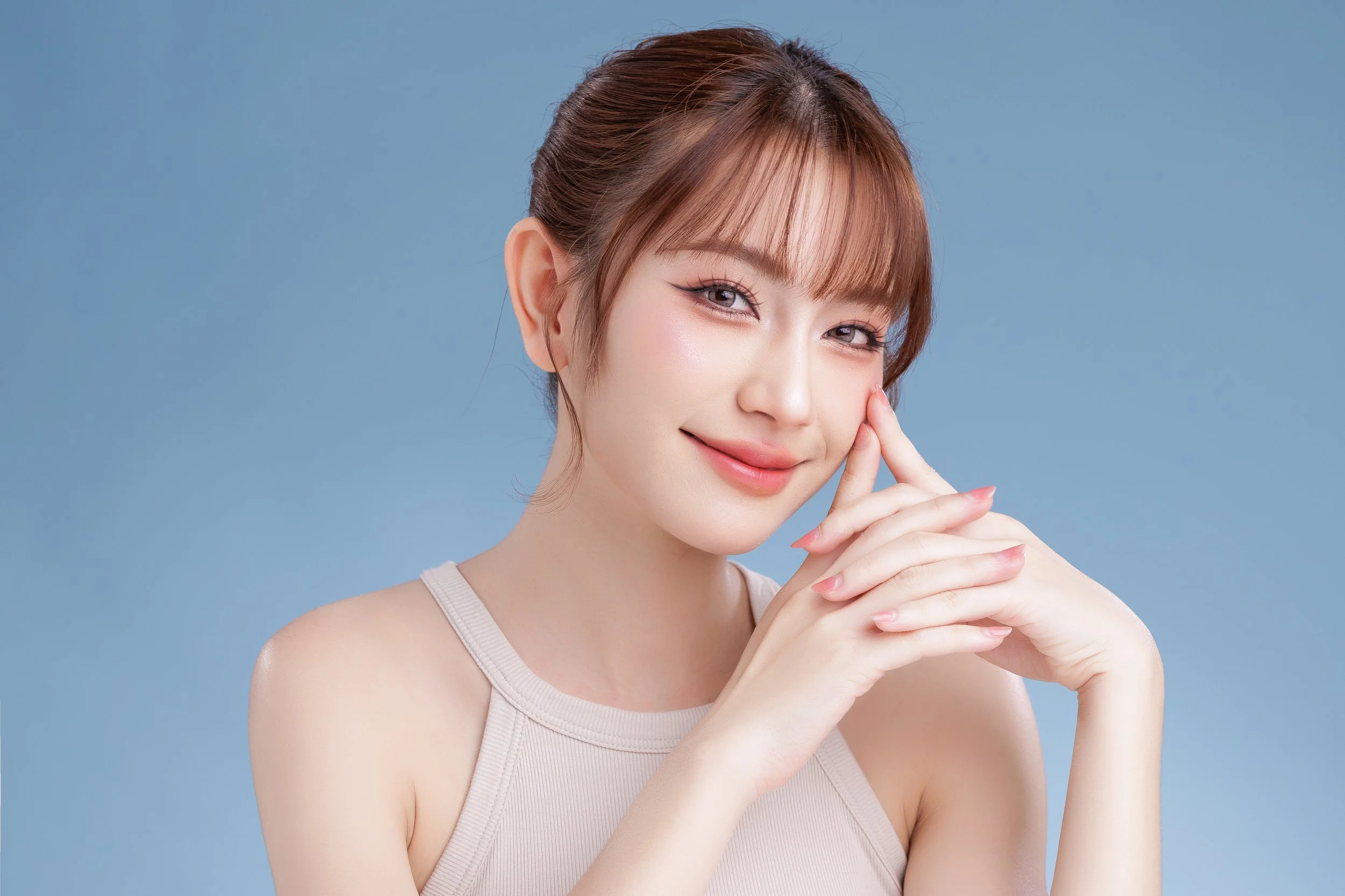 A young woman with light brown hair styled in a bun, smiling gently with her hands clasped near her face, wearing a beige sleeveless top against a light blue background.
