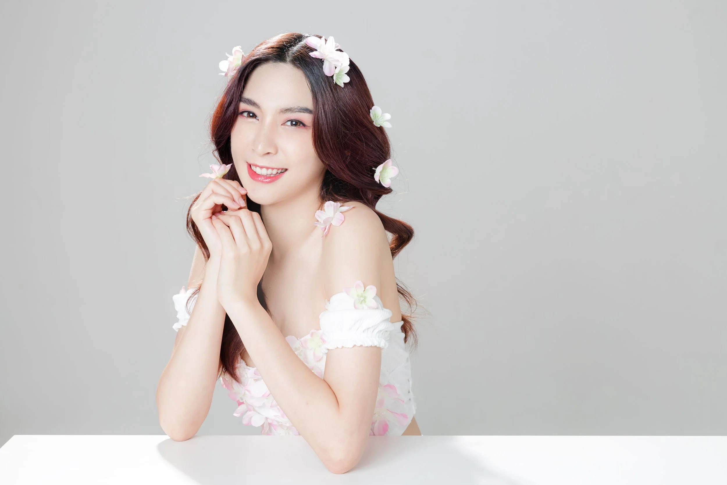 A smiling young woman with long, wavy brown hair with pink tint, decorated with pink and white flowers, sitting at a white table against a plain gray background. She is wearing a white off-the-shoulder dress with floral embellishments and has her han