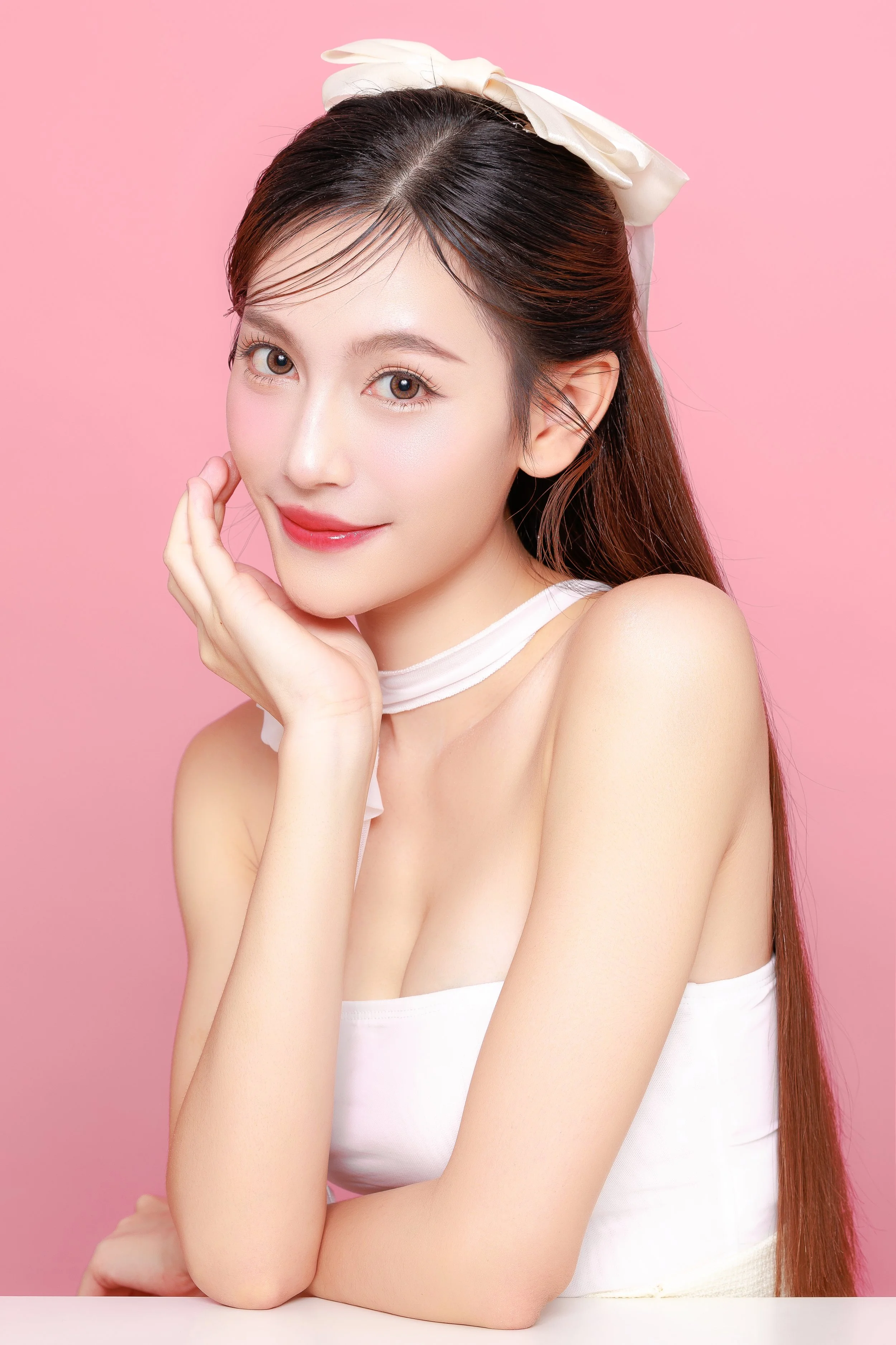 A woman with long dark hair, wearing a white ribbon in her hair and a white strapless top, smiling softly with her hand resting on her cheek against a pink background.