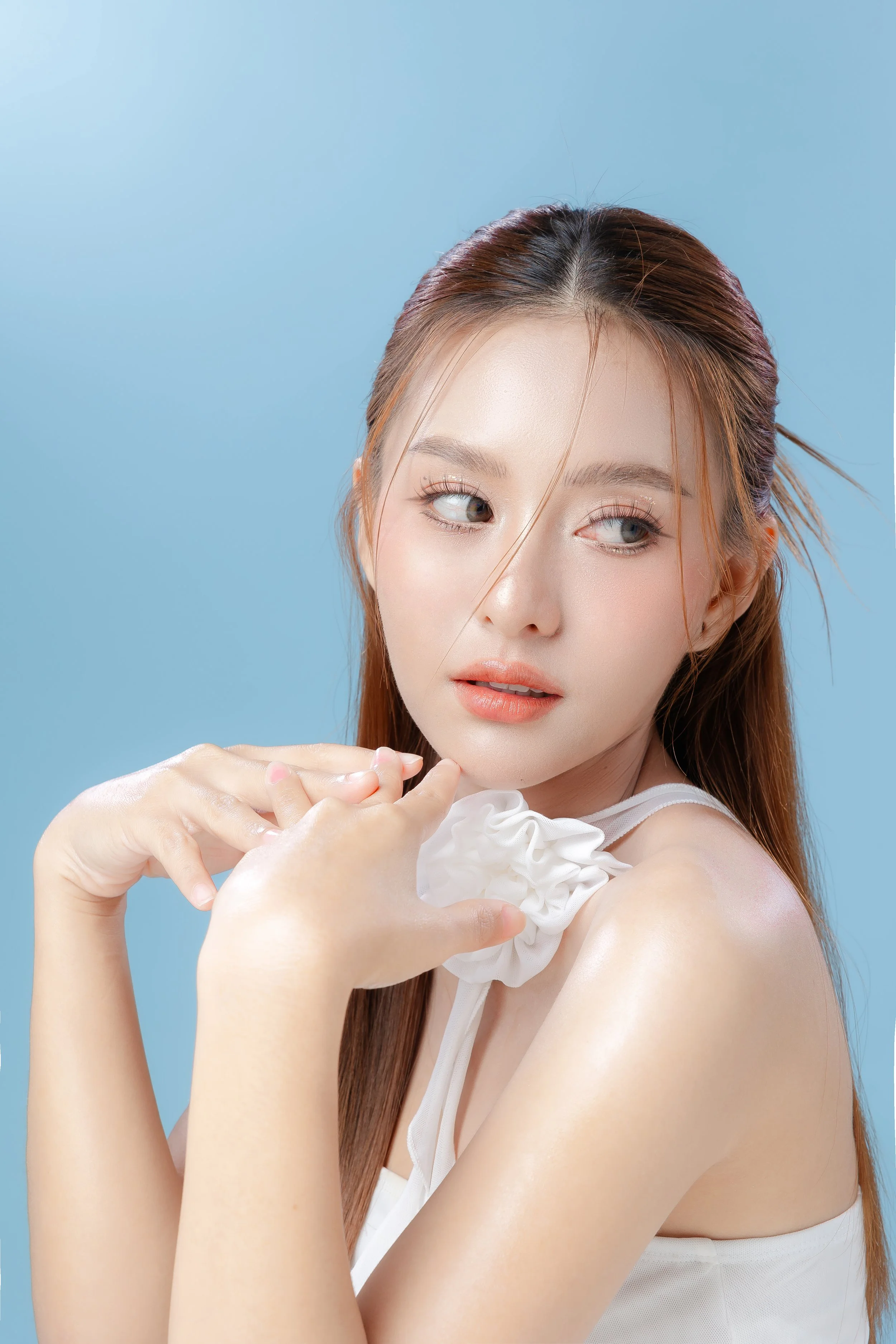A young woman with light skin and long light brown hair styled with a few loose strands, posing against a light blue background. She is wearing a white top with a ruffled shoulder detail and looking to her left with her hands gently touching her chin