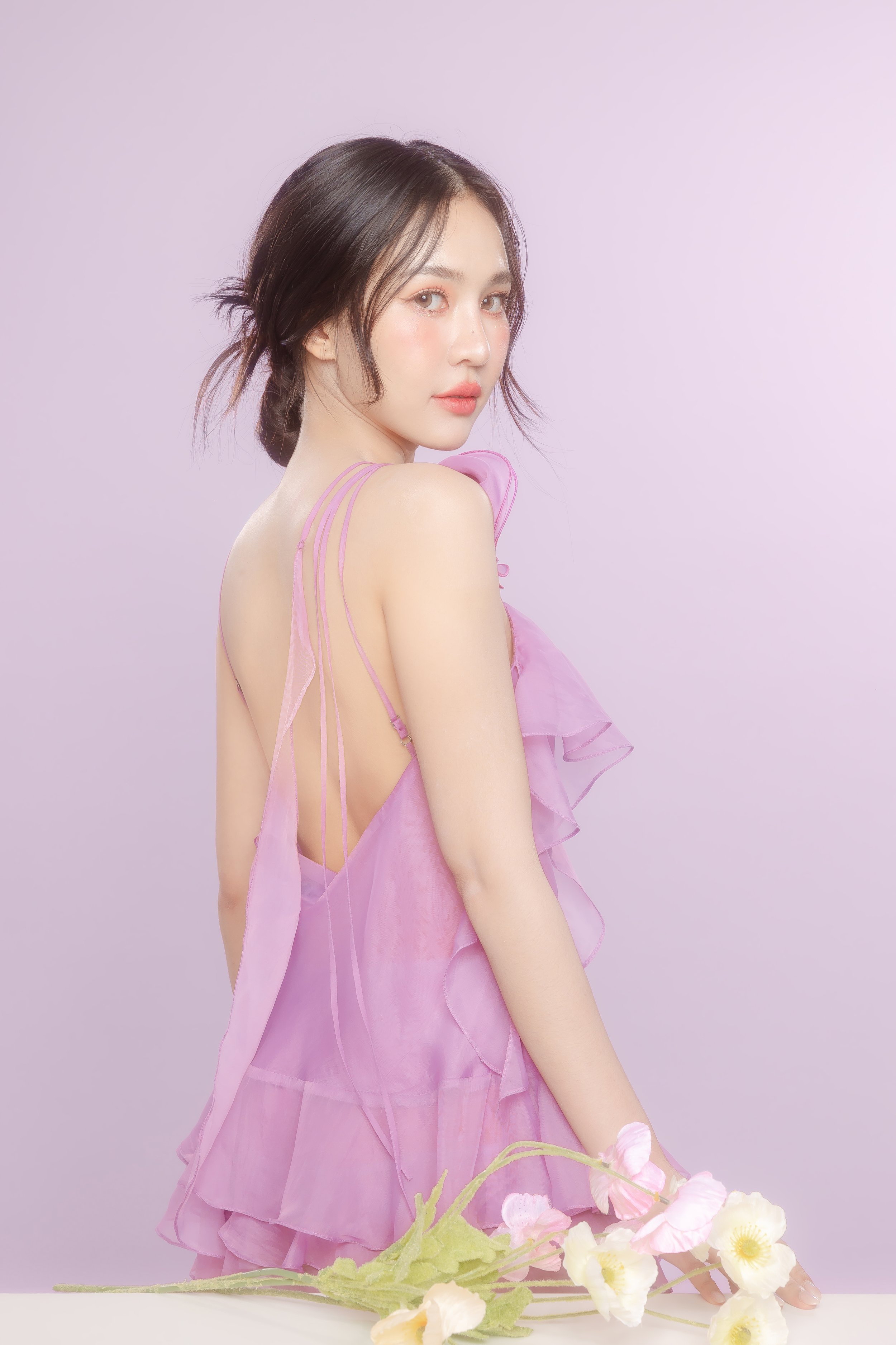 Young woman with dark hair styled in a loose, low bun, wearing a pink, ruffled, sheer dress, standing against a light purple background with pink and white flowers on the table in front of her.