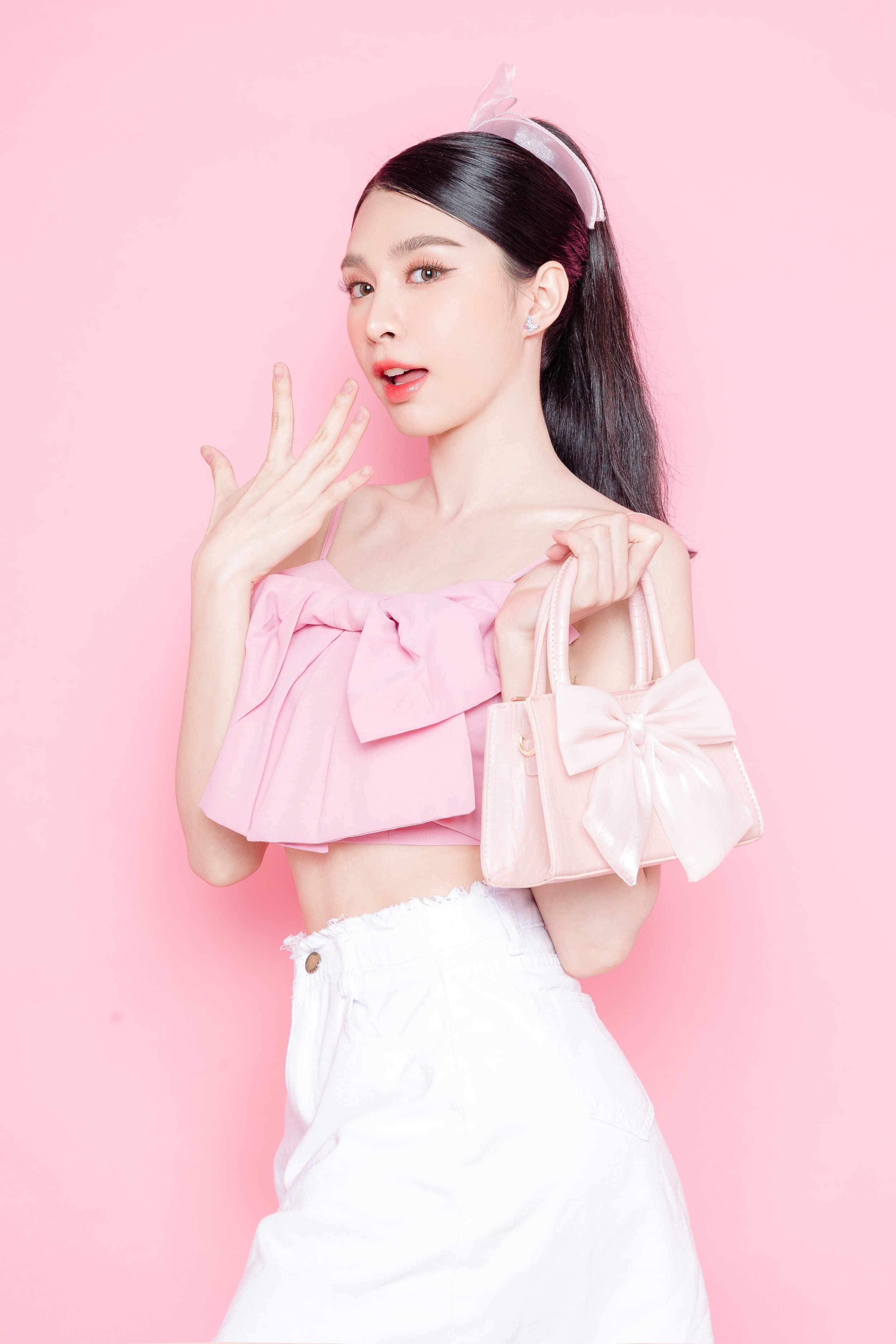 A young woman with long black hair, wearing a pink top with a large bow and white high-waisted pants, posing against a pink background with a pink handbag and a pink headband.