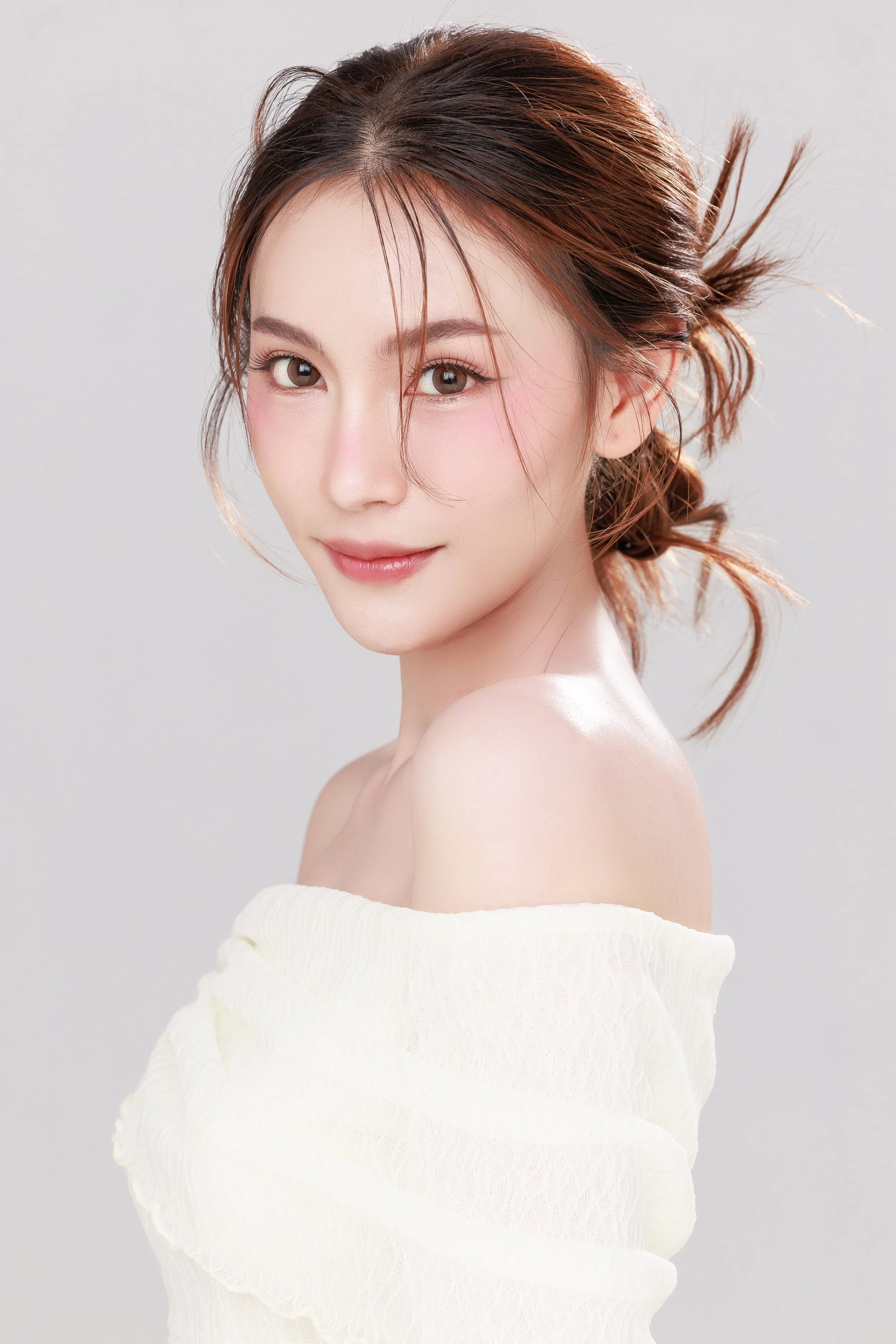 A young woman with brown hair styled in loose, messy waves, wearing a white off-shoulder top, looking at the camera with a slight smile against a plain light background.