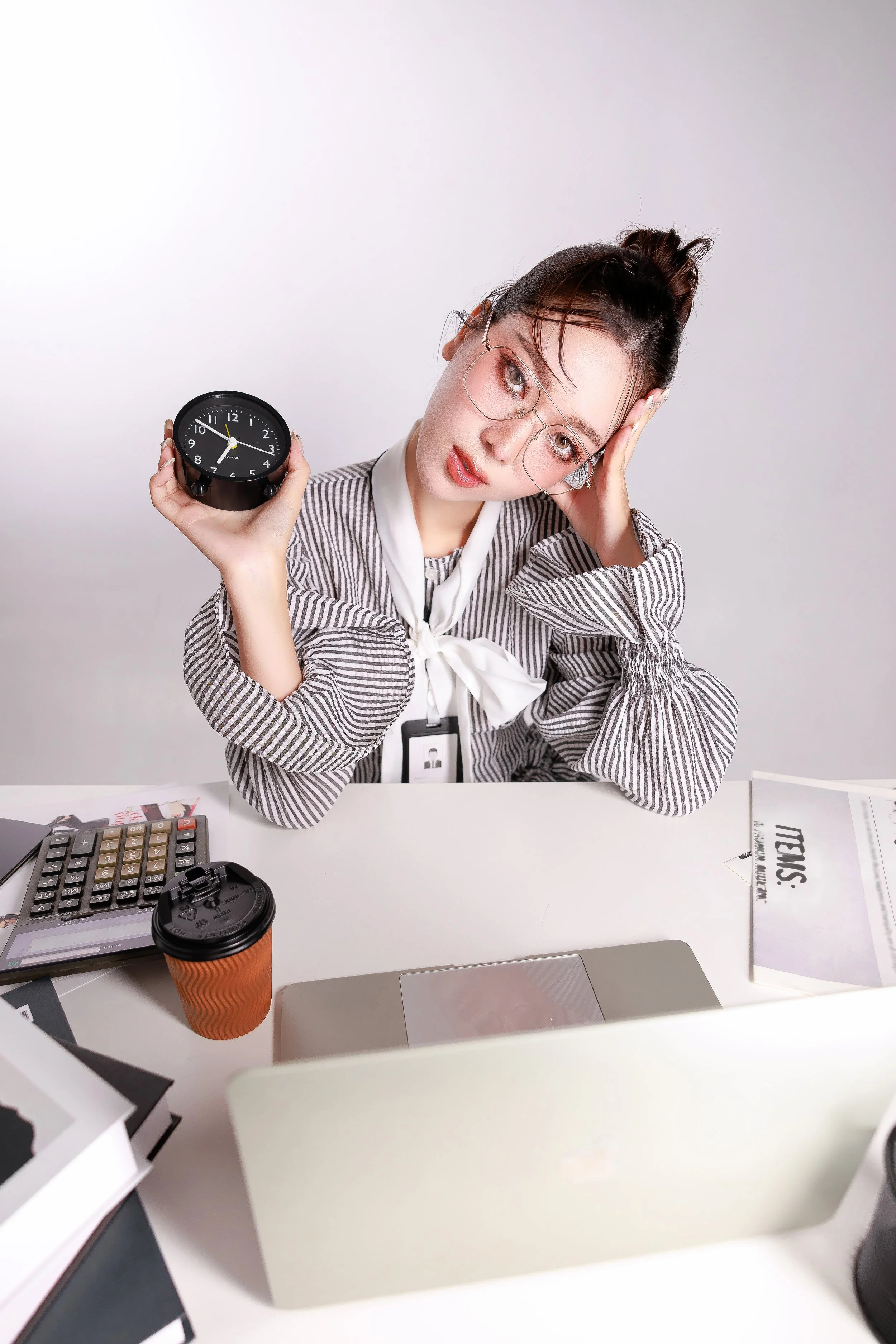 Disoriented woman in glasses holding a black alarm clock showing 9:10, sitting at a cluttered desk with a coffee cup, calculator, laptop, and papers, looking stressed or tired.