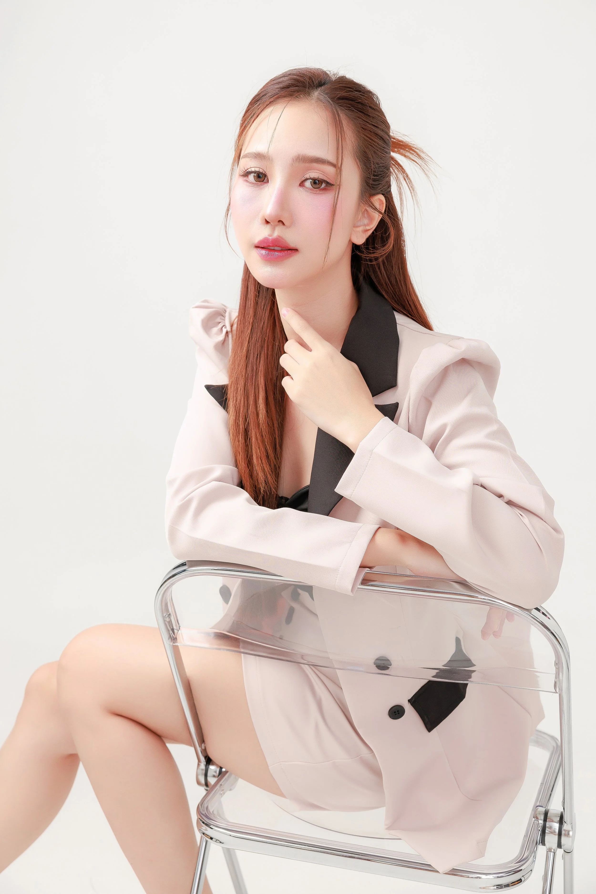 A woman with red hair sitting on a transparent chair, wearing a pink blazer with black accents, against a plain white background.