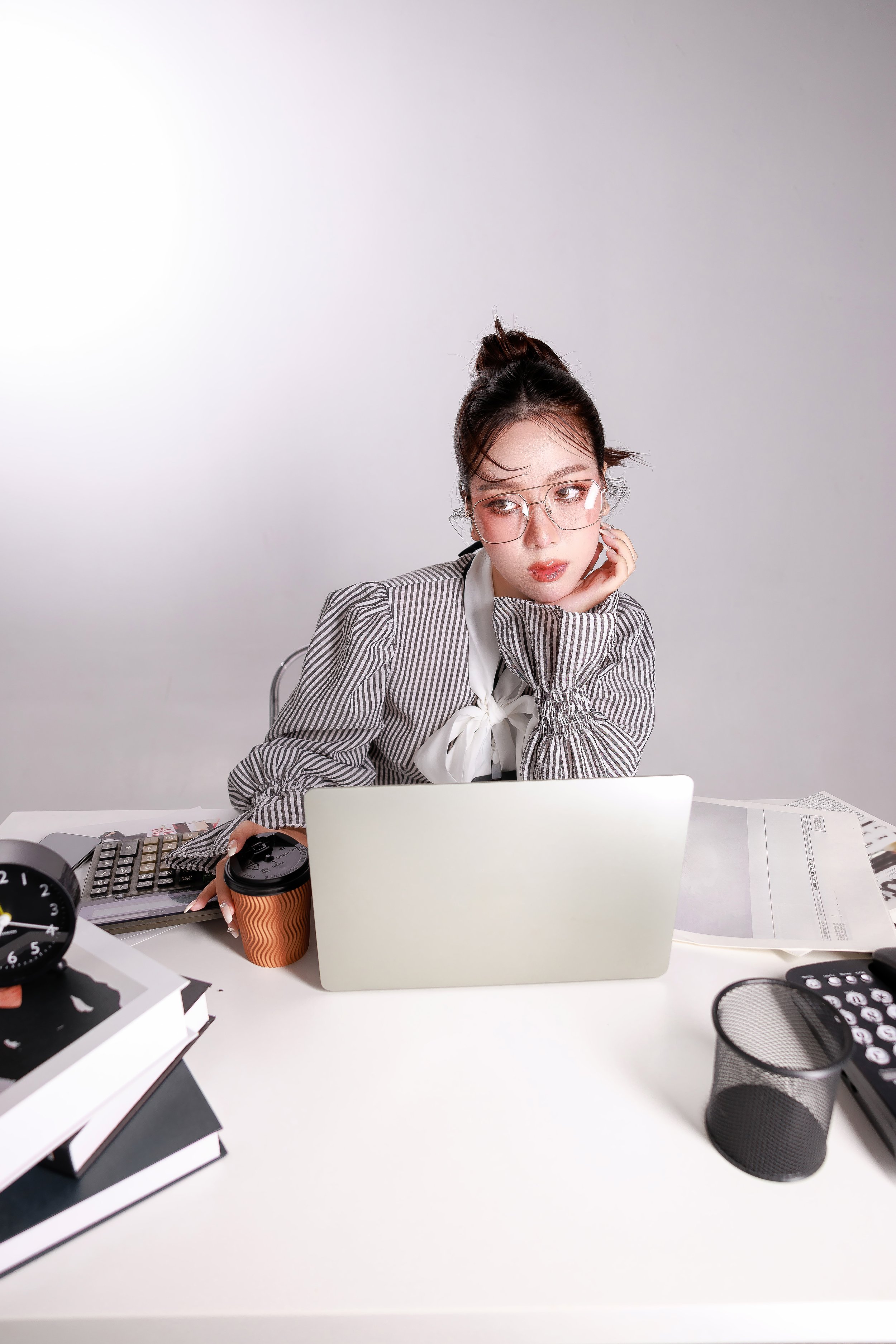 A young woman with glasses and a striped blazer sitting at a cluttered desk, looking to the side while working on a laptop, surrounded by papers, books, a calculator, clock, coffee cup, and office supplies.