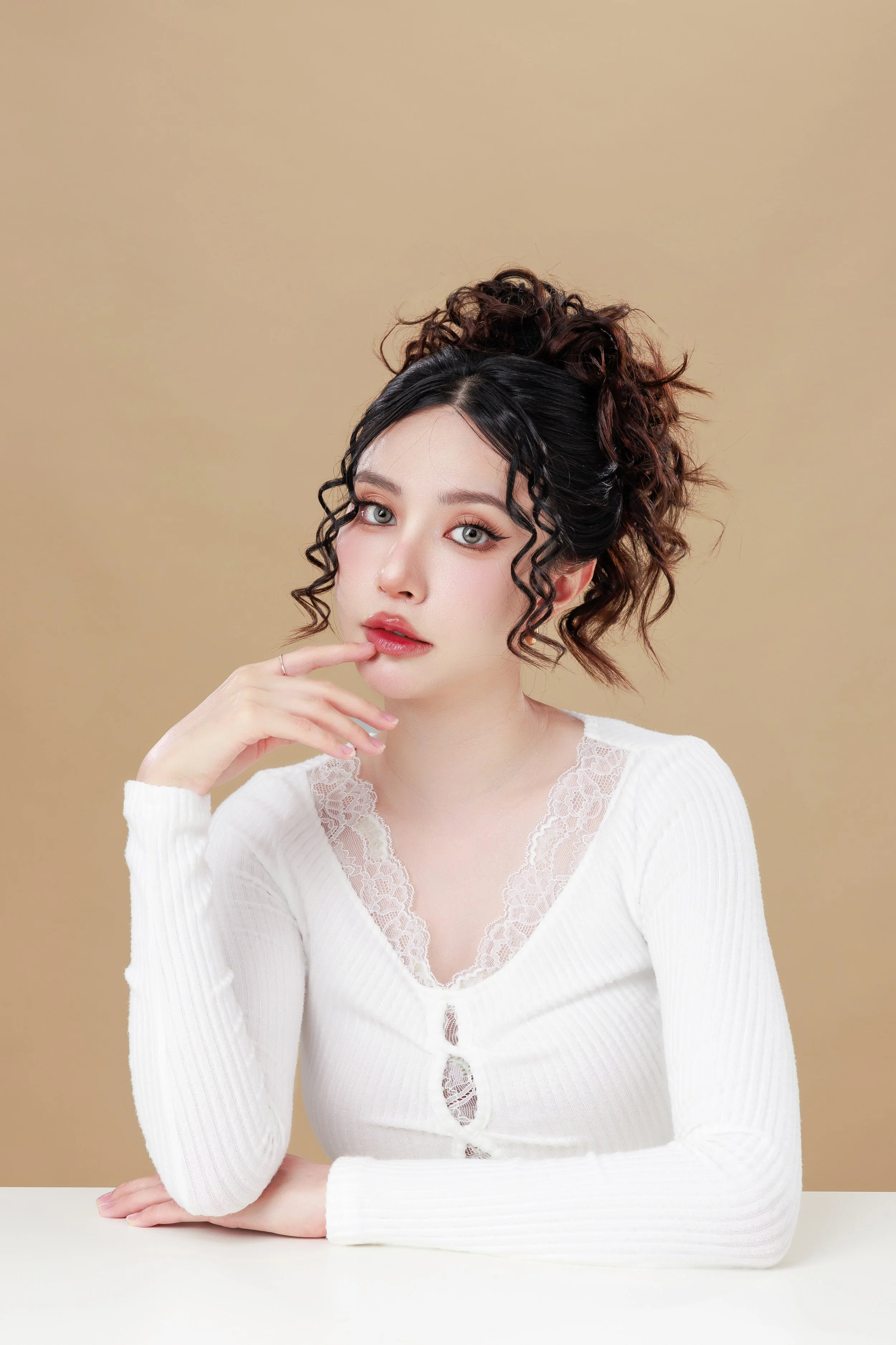 A young woman with curly dark hair styled in a high updo, wearing a white long-sleeve top with lace details, sitting at a white table against a beige background, with her left hand resting on her face.