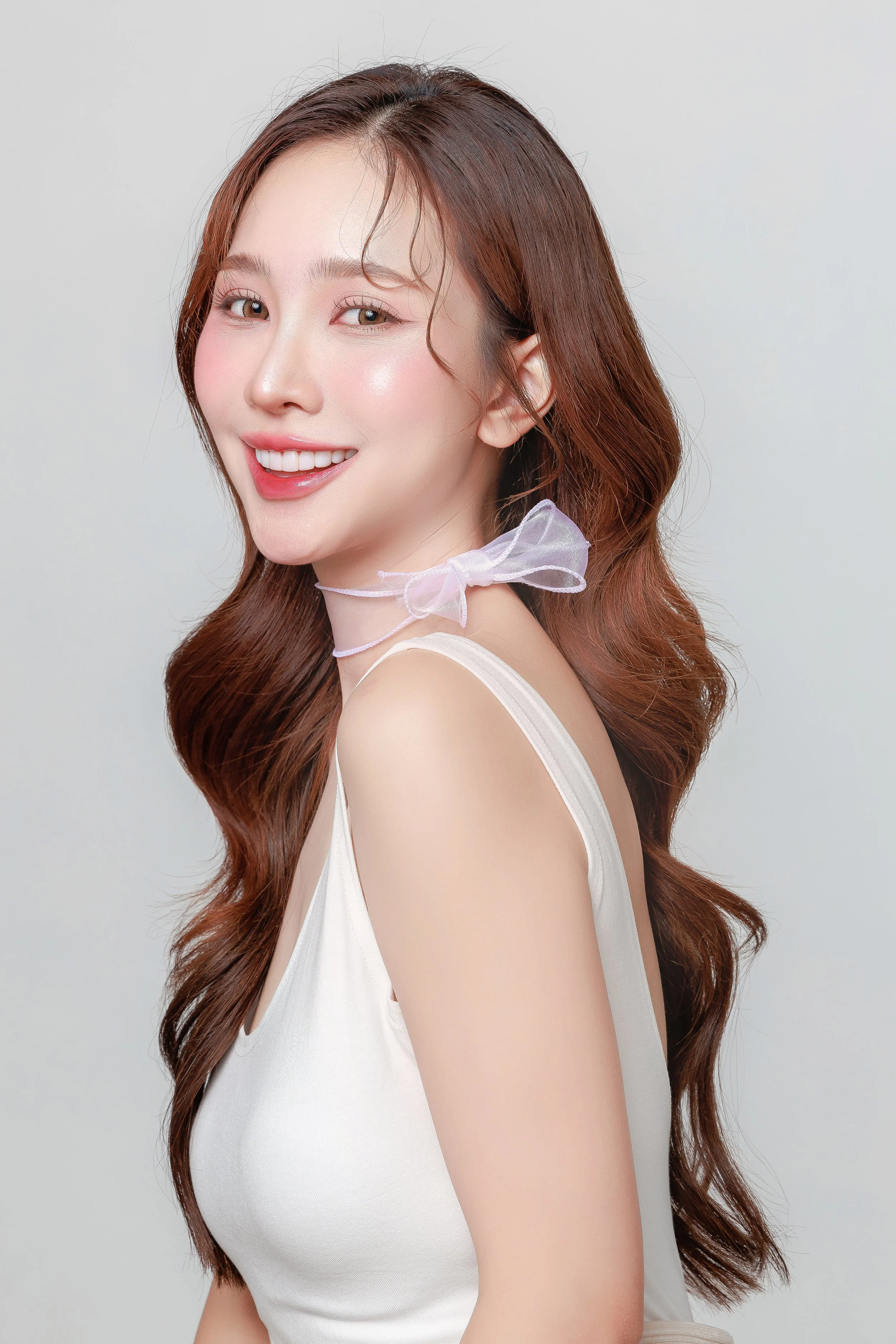 A woman with long, wavy auburn hair is smiling at the camera. She is wearing a sleeveless white top and a sheer, light pink ribbon choker tied in a bow around her neck. The background is plain and light-colored.