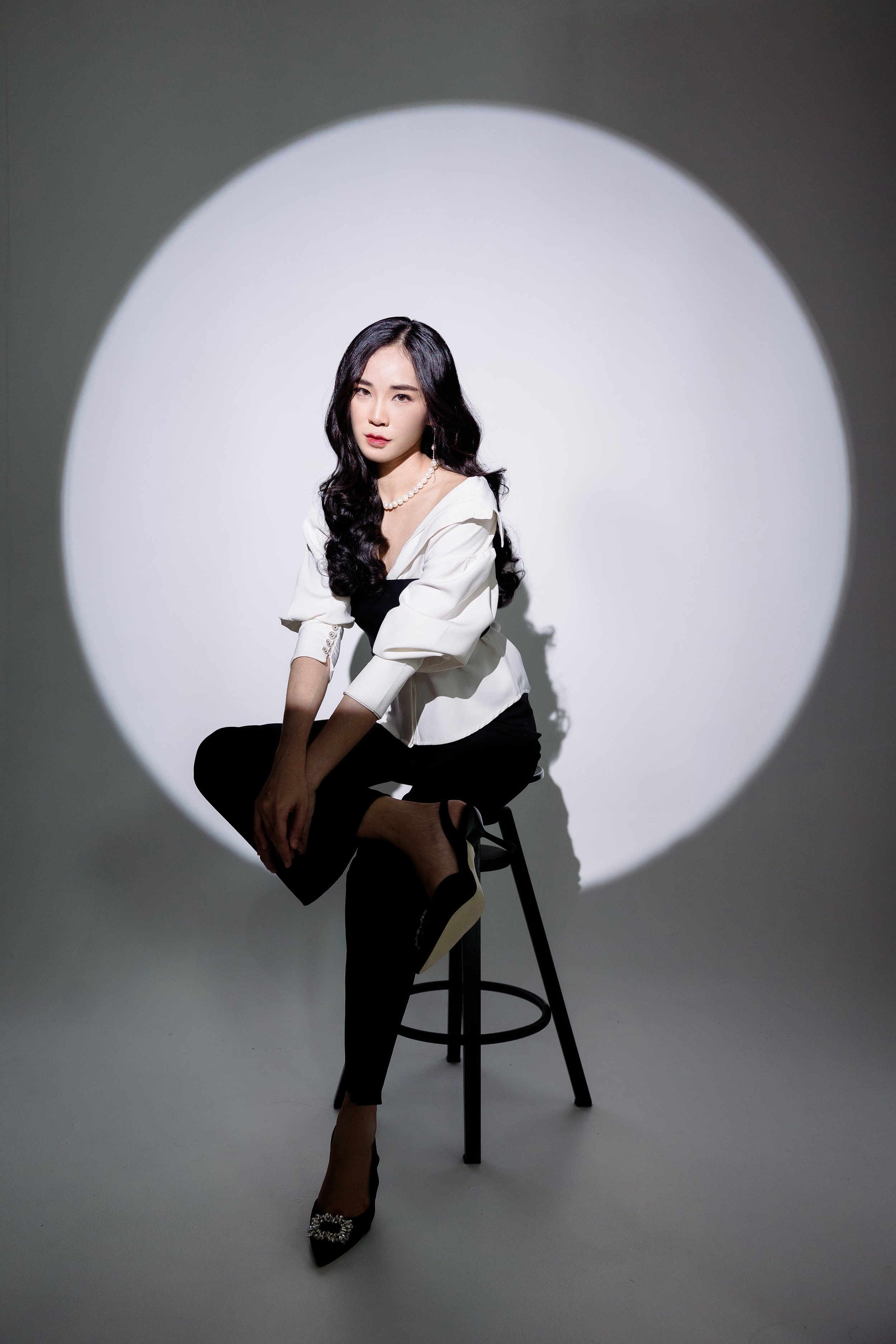 A woman with long black wavy hair sitting on a black stool, wearing black pants, a white blouse, a pearl necklace, and black high heels, with a spotlight creating a circular light pattern on the plain gray background.