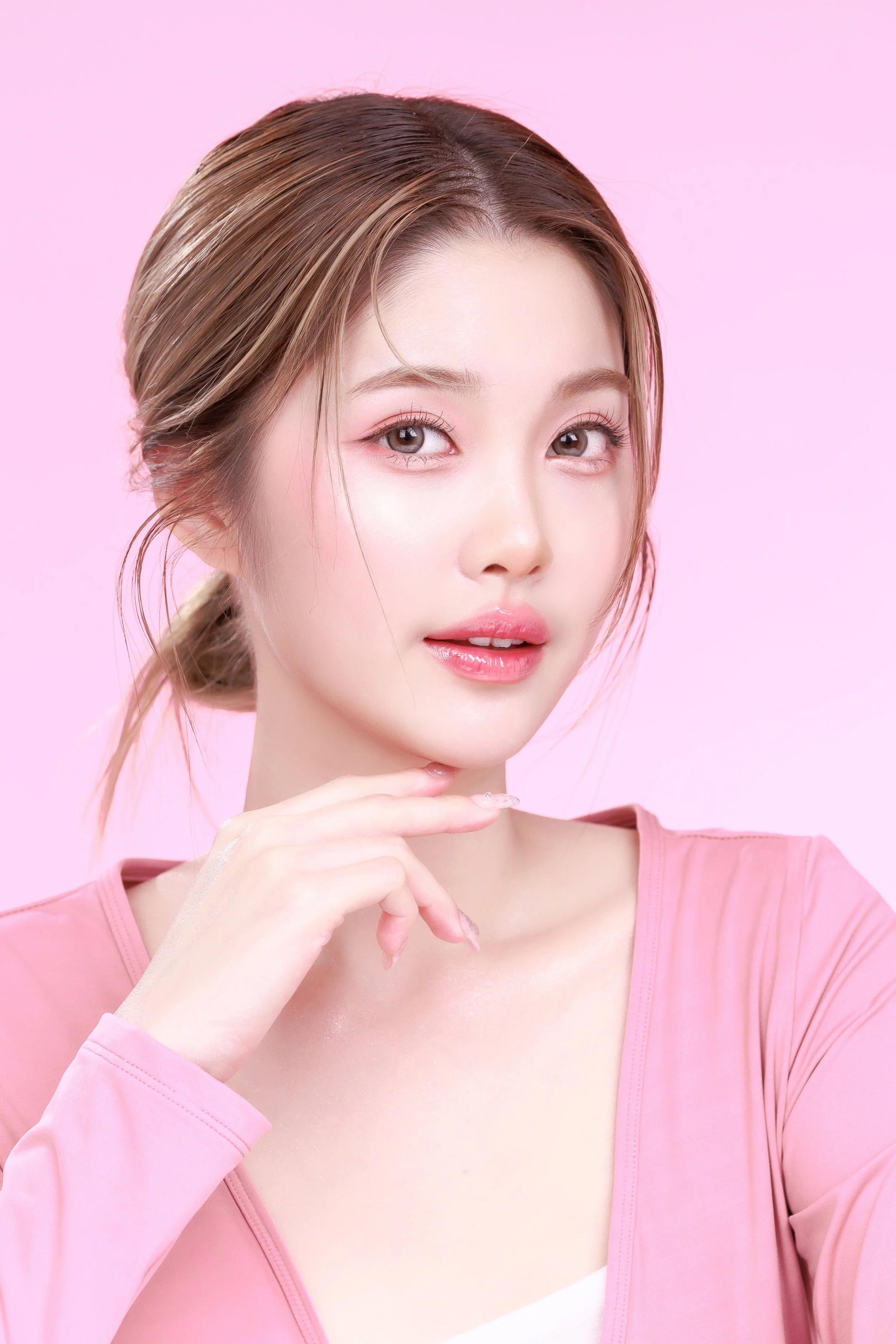 A young woman with long, light brown hair styled in a low bun, wearing a pink top, and posing against a pink background. She has natural makeup with pink lipstick and light eyeshadow.