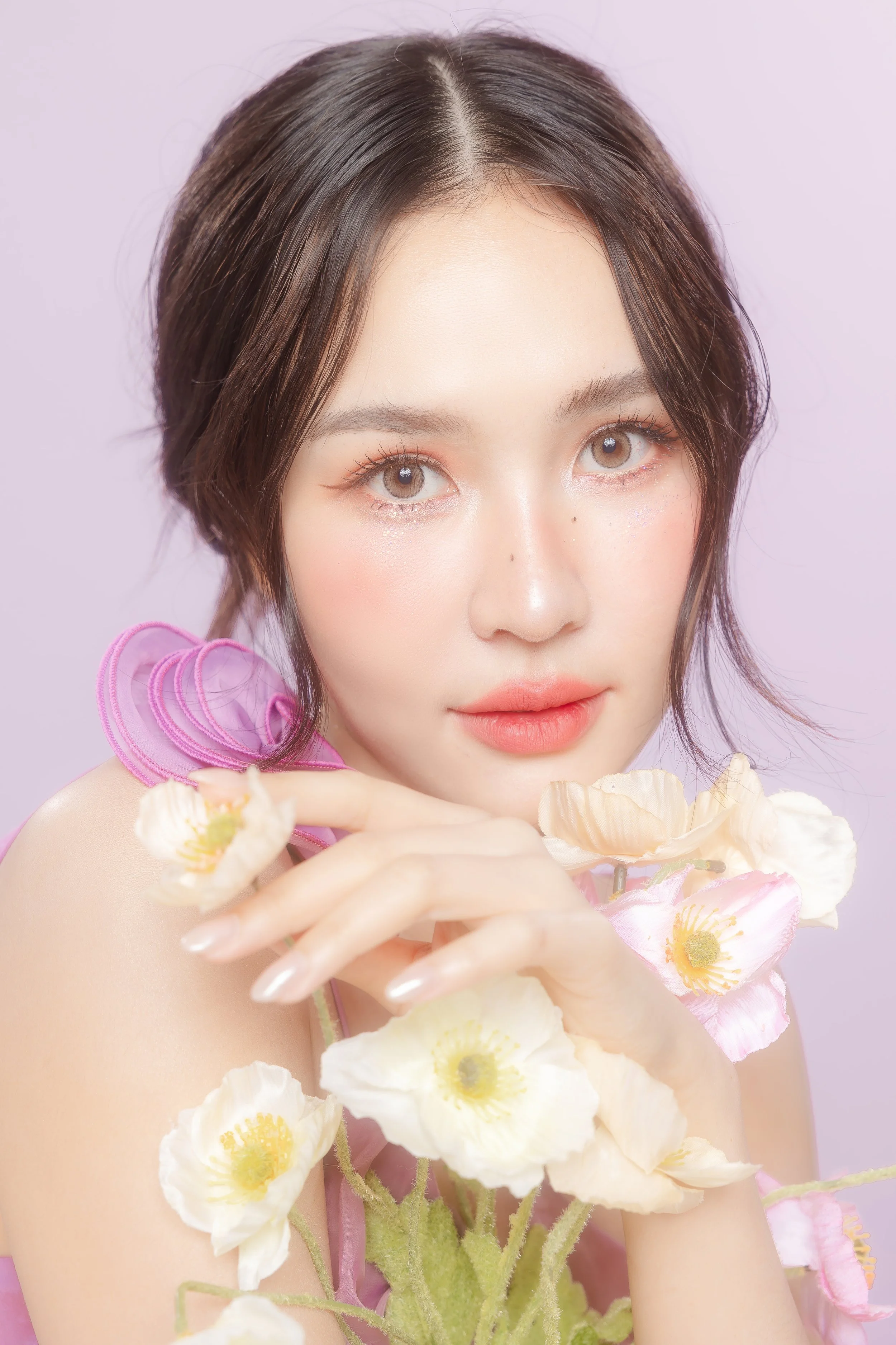 Portrait of a young woman with short dark hair, wearing pink flowers and holding white and pink flowers, against a pastel purple background.