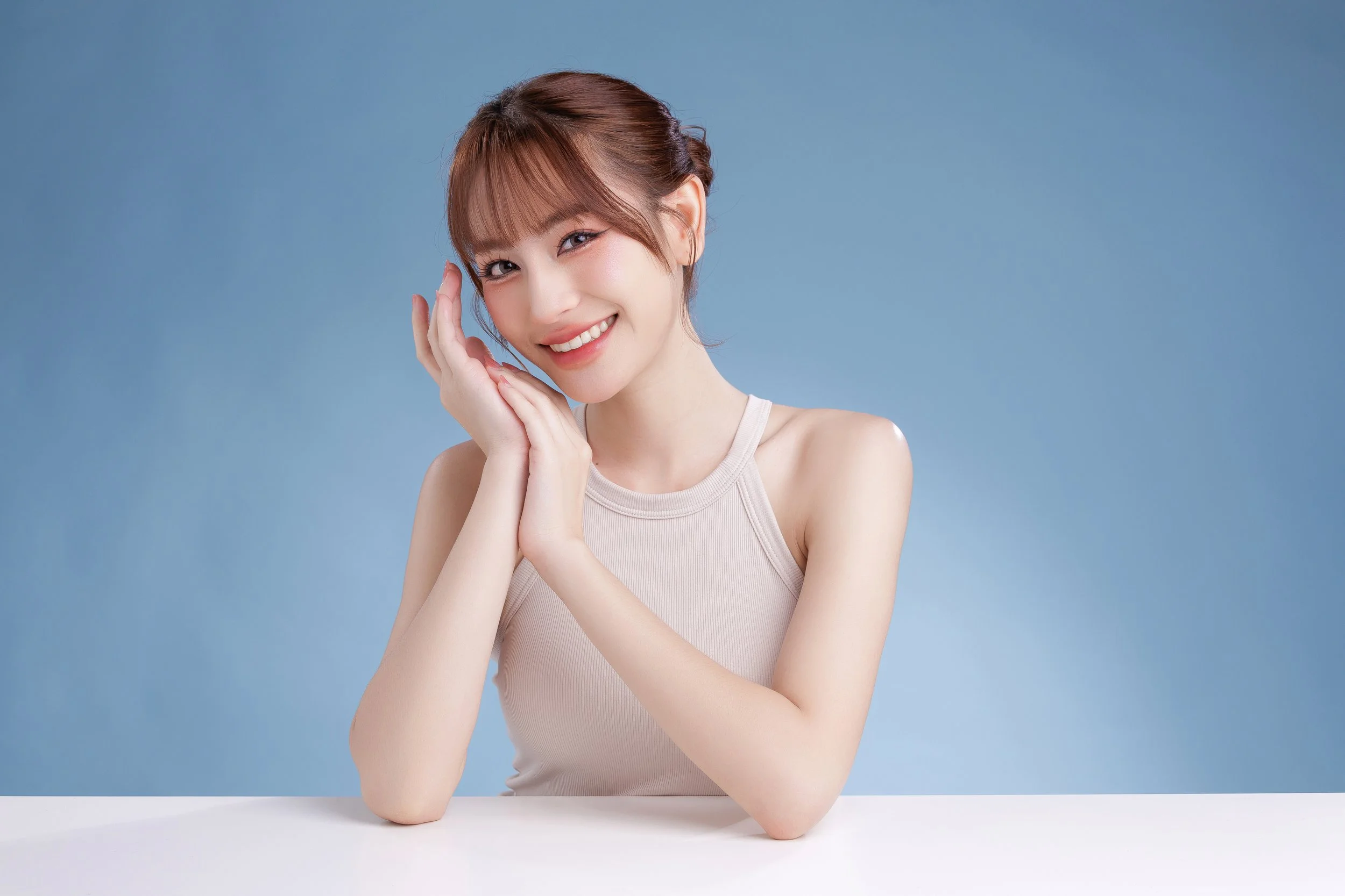 A young woman with brown hair styled in a bun, smiling and touching her face with both hands, wearing a sleeveless beige top against a blue background.