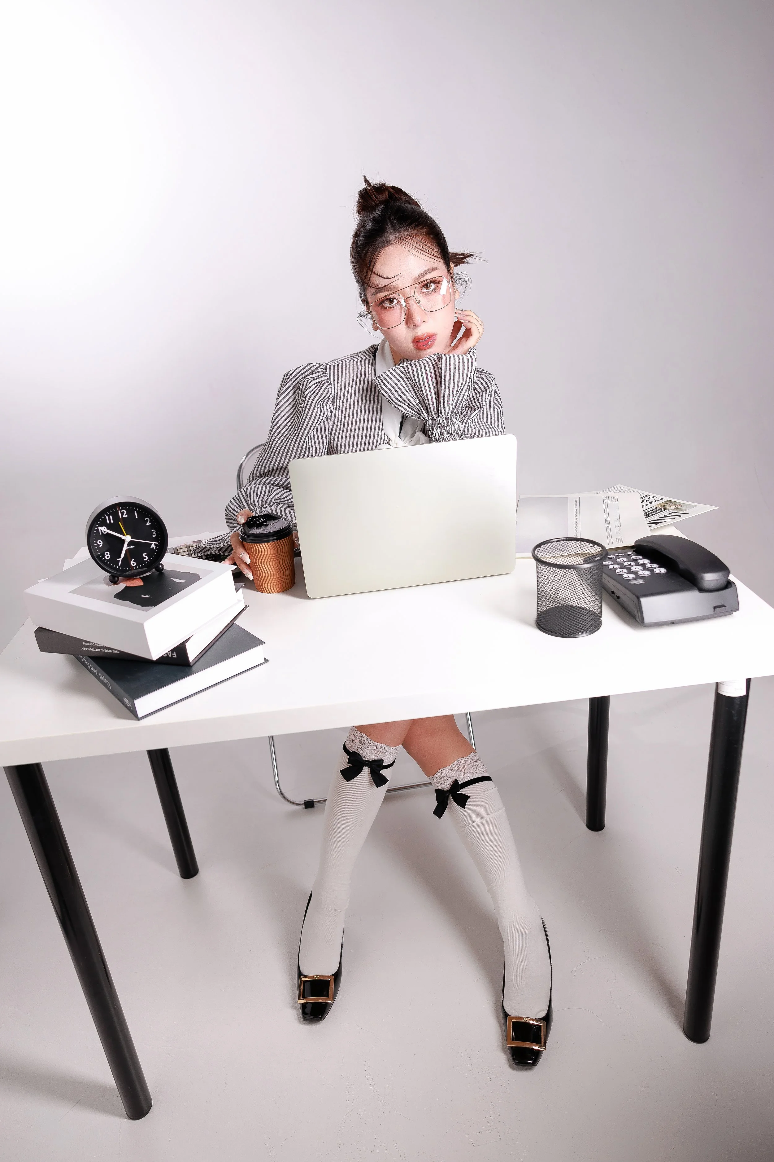 A young woman sitting at a white desk in a modern office setting. She has her hand resting on her face, wears glasses, a striped blazer, and knee-high socks with bows and heels. She is surrounded by office supplies including a clock, a coffee cup, a 