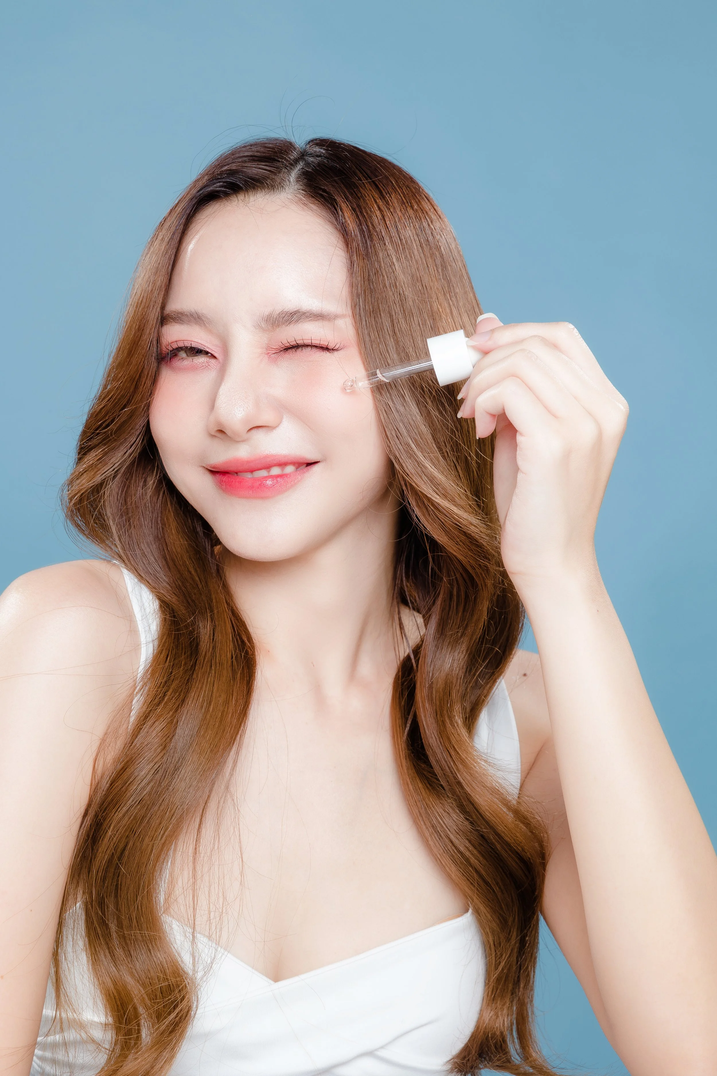 Young woman with long, wavy brown hair smiling with eyes closed, holding a dropper with skincare serum near her face against a solid blue background.