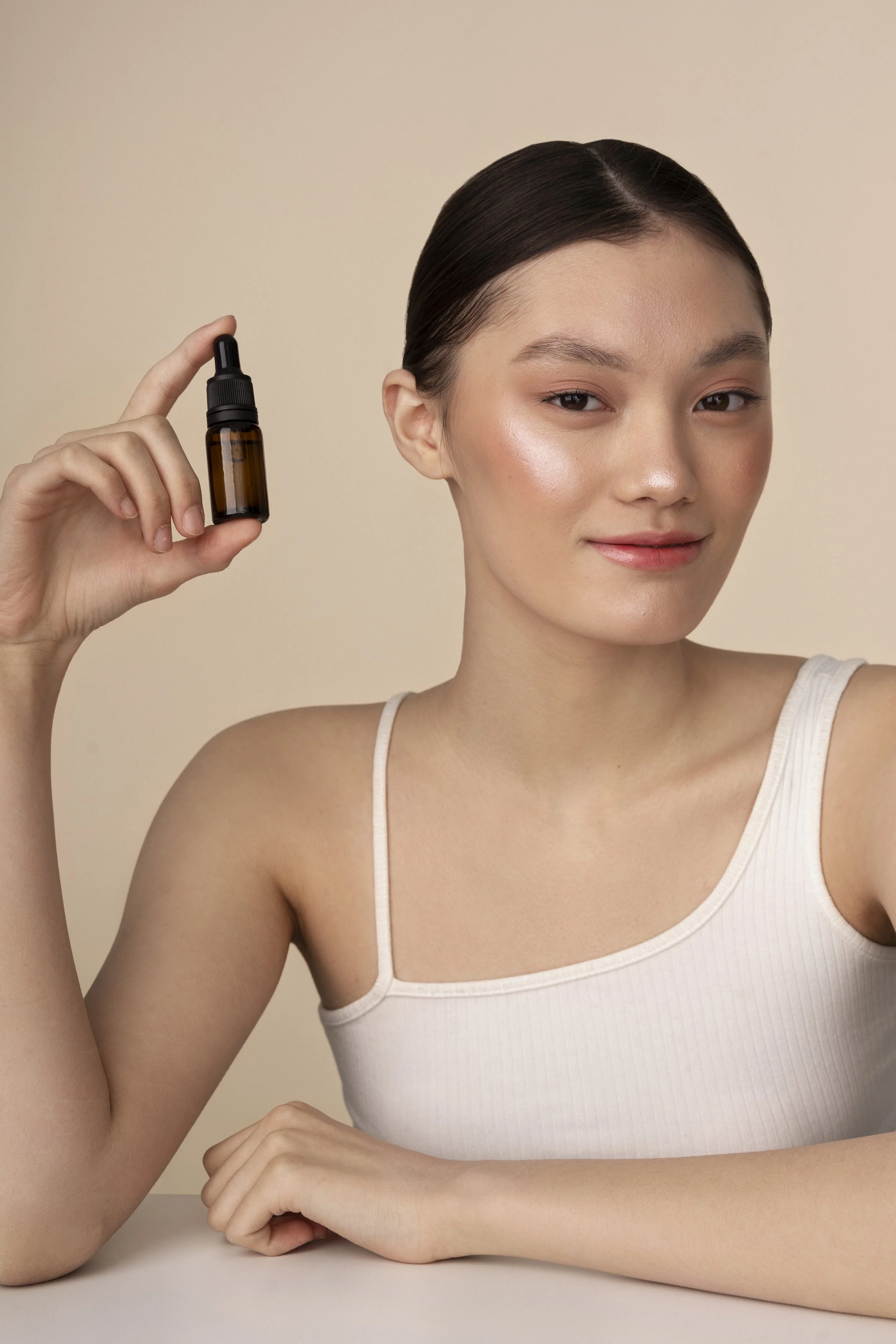A young woman with clear skin and dark hair in a bun holds a small brown glass dropper bottle near her face against a beige background.
