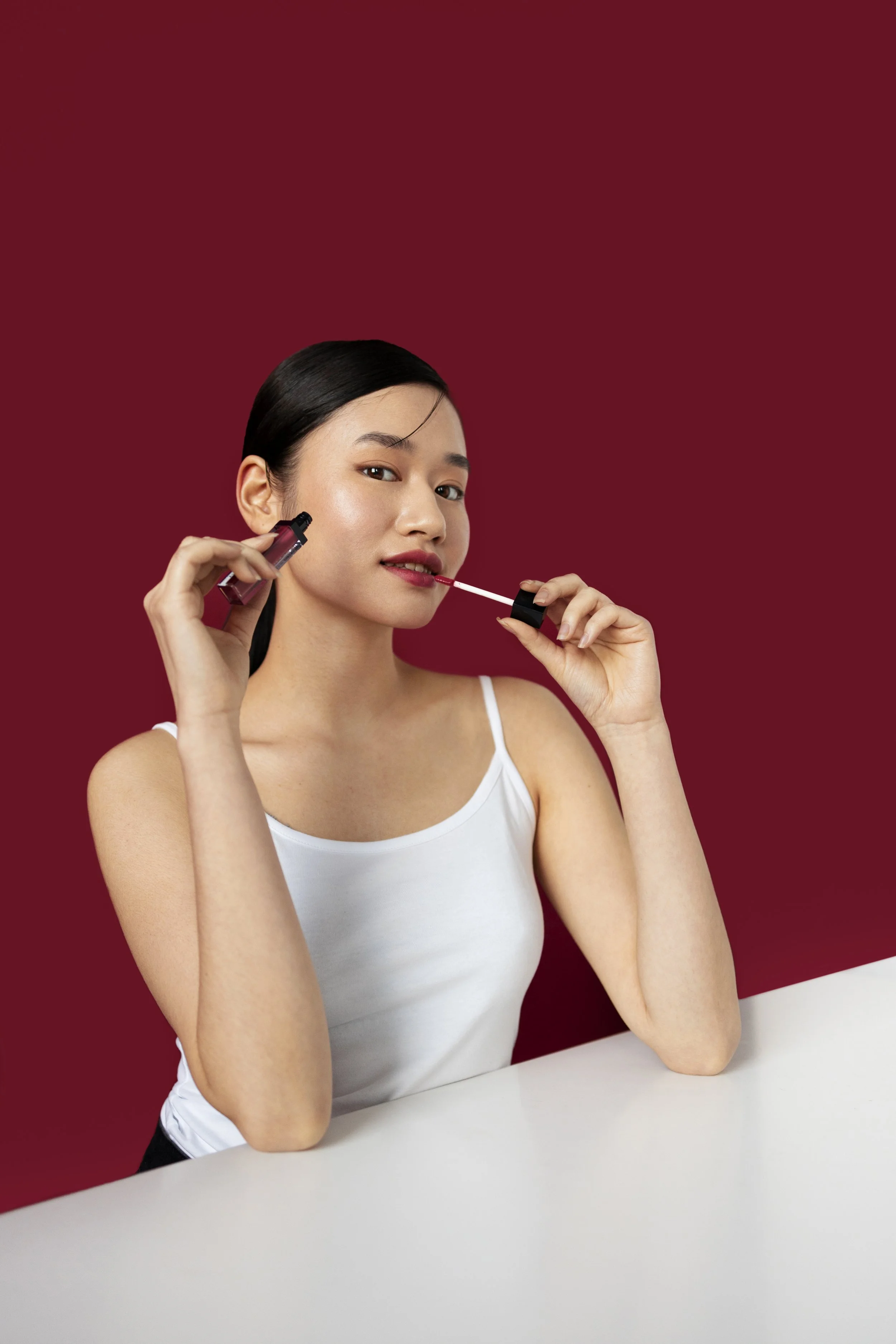 A woman with black hair applying red lipstick in front of a red background.