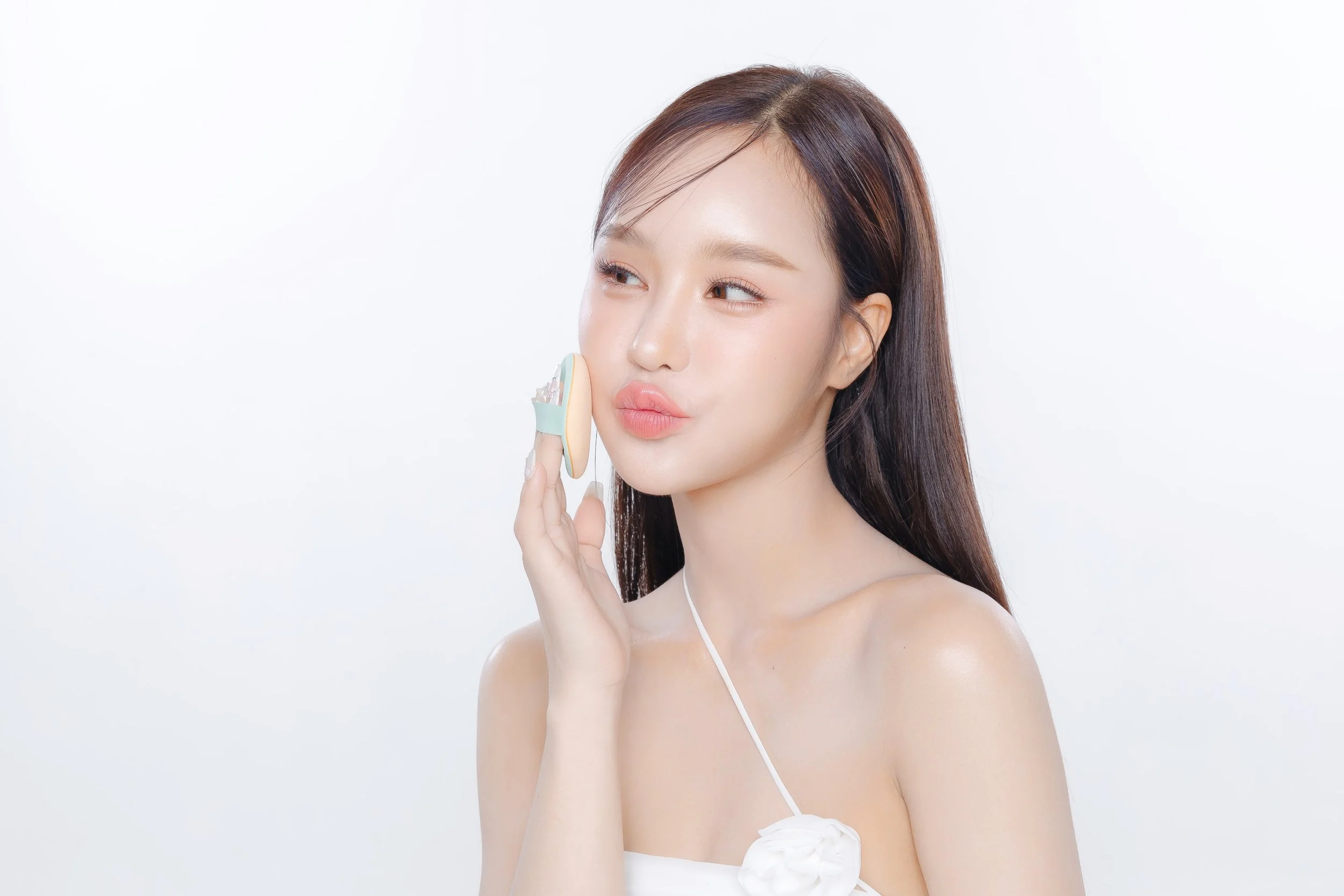 A young woman with long brown hair holding a pastel-colored makeup sponge to her cheek, facing slightly to the side with a soft expression and lips pursed, against a plain white background.