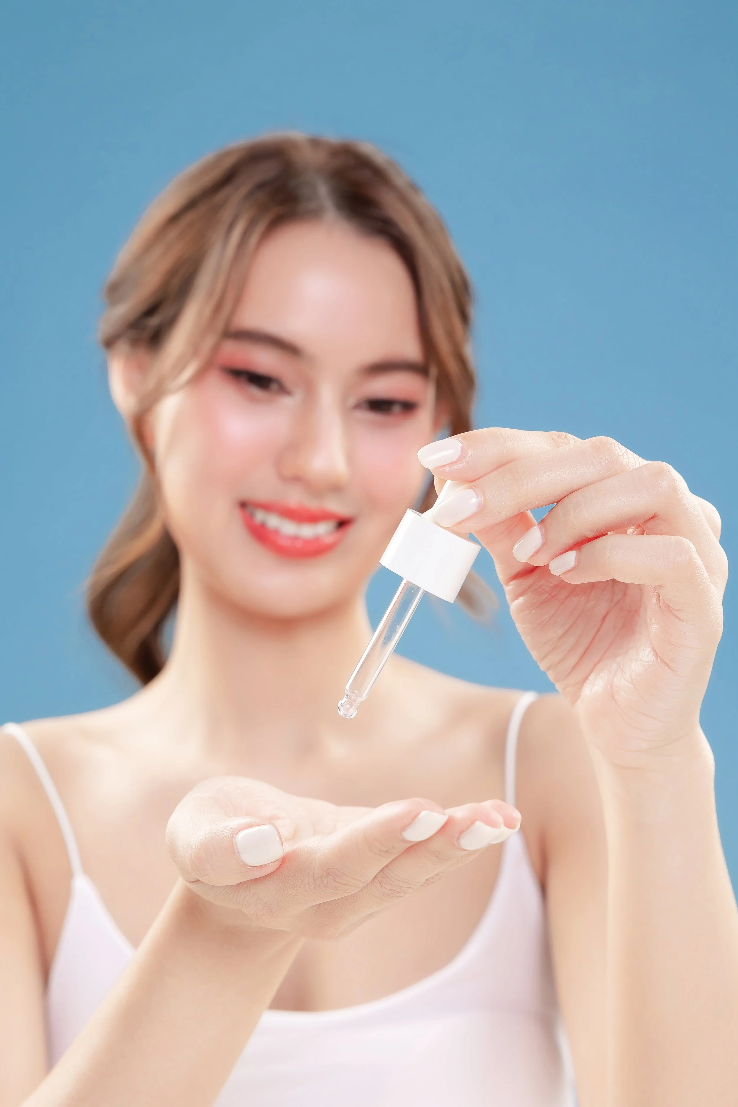 A woman holding a dropper and applying skincare or serum to her hand against a blue background.