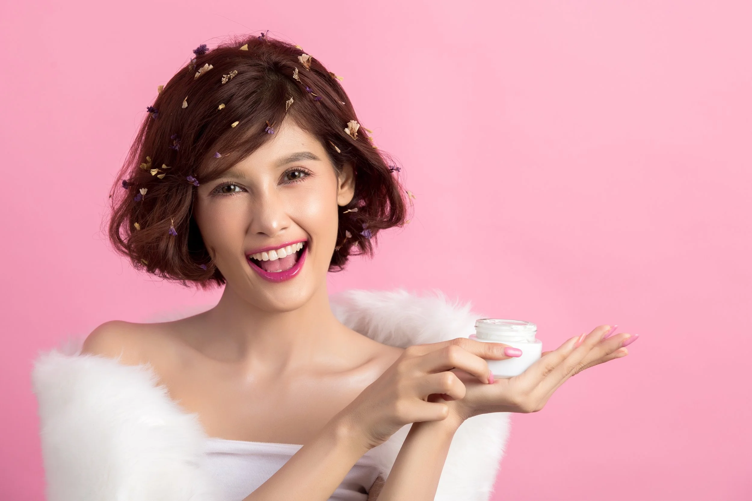 A woman with short brown hair, wearing a white top and a white furry shawl, holding a jar of cream, smiling with pink lipstick, against a pink background.