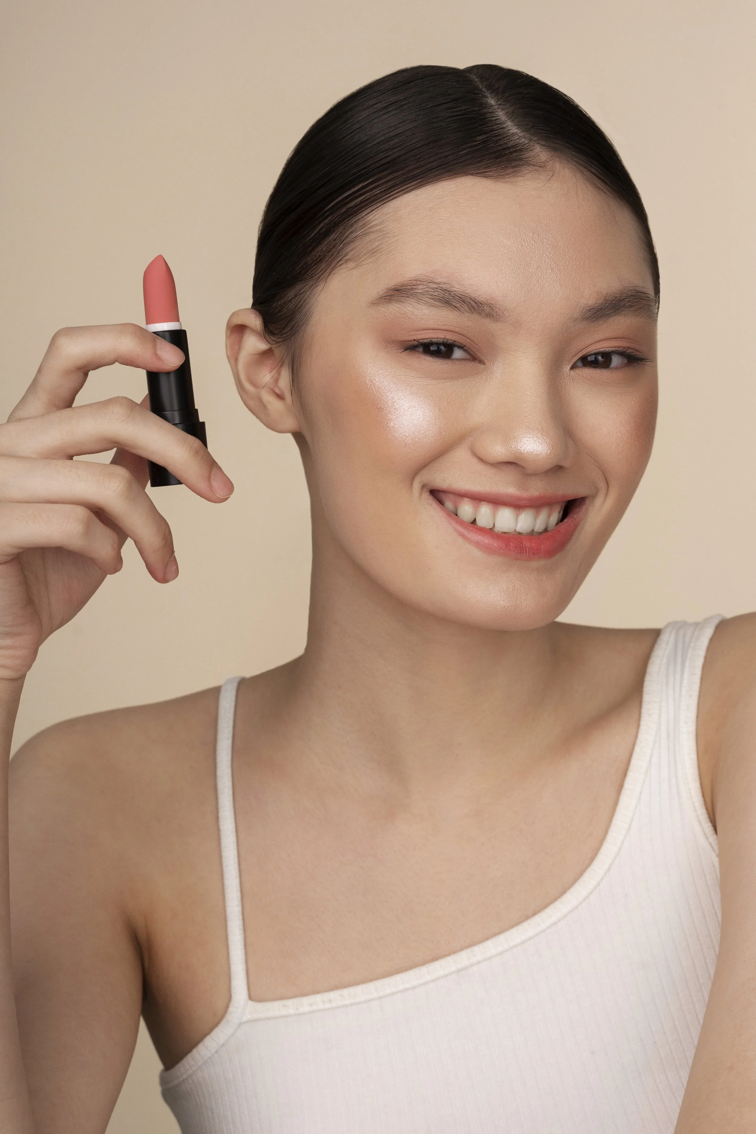 A woman with dark, pulled-back hair smiling while holding a pink lipstick near her face.