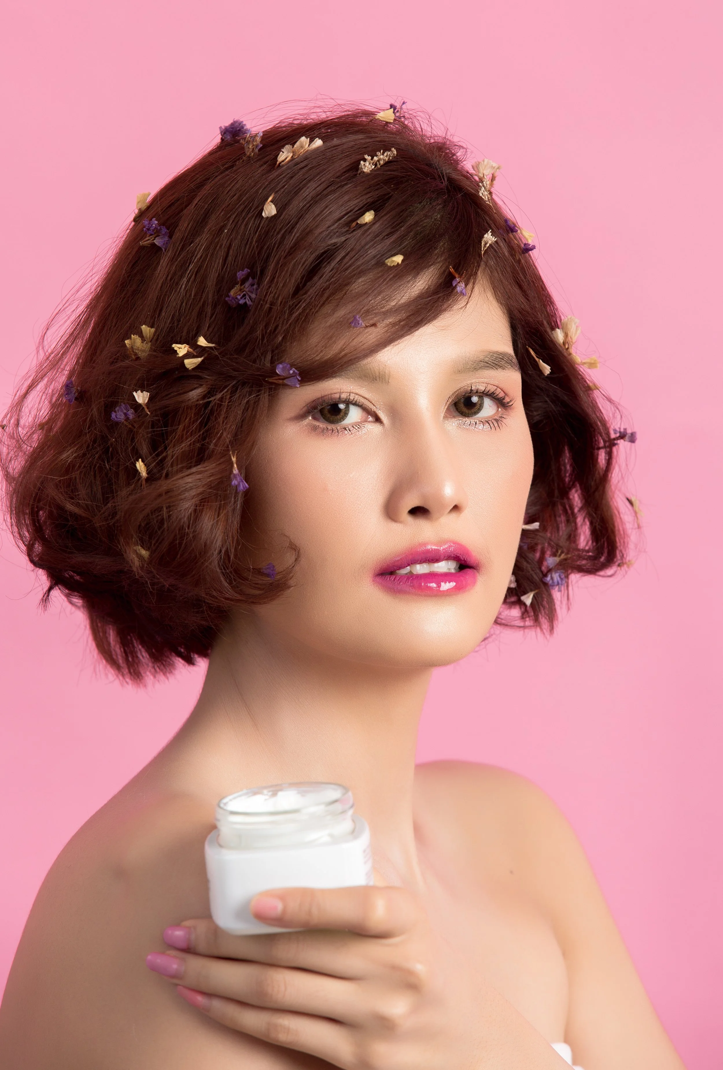 Young woman with short, wavy brown hair decorated with small purple and yellow flowers, holding a jar of cream against a pink background.