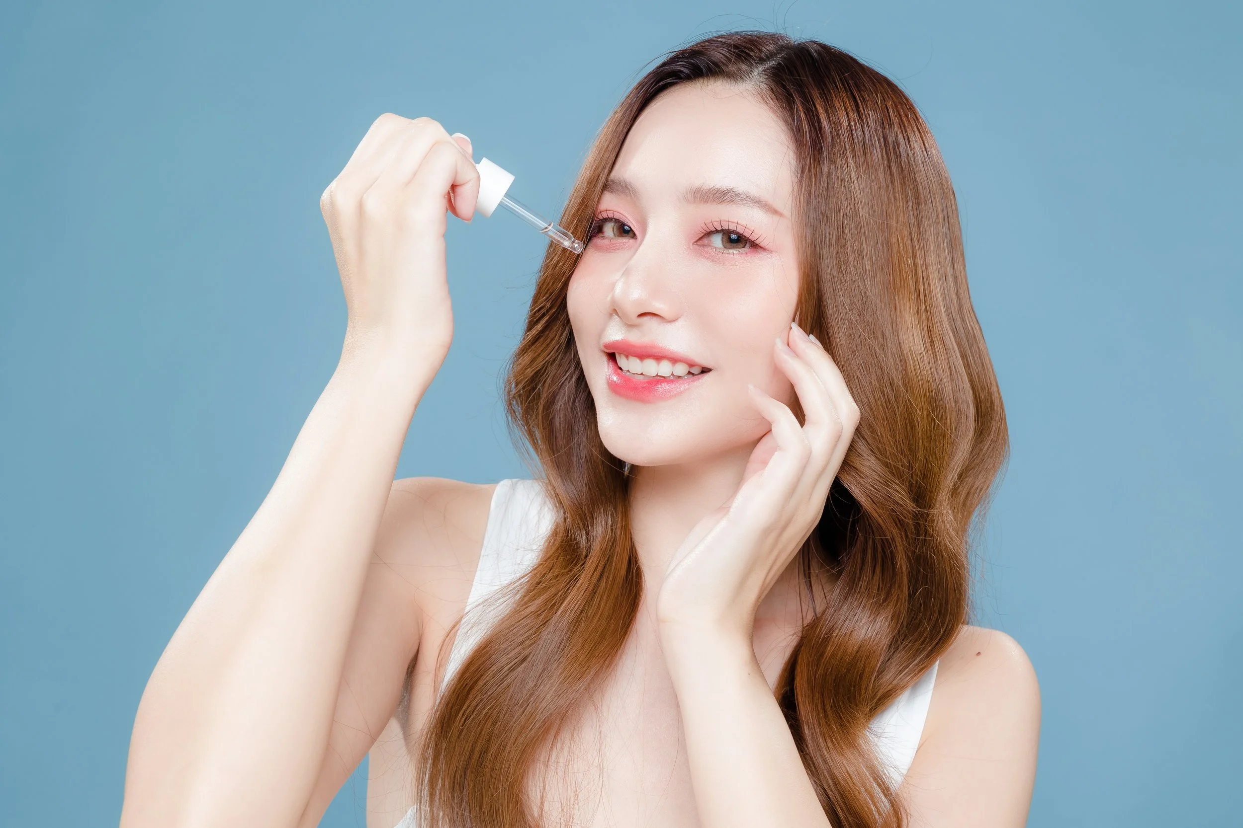 A woman with long, wavy brown hair applies skincare using a dropper against a blue background.