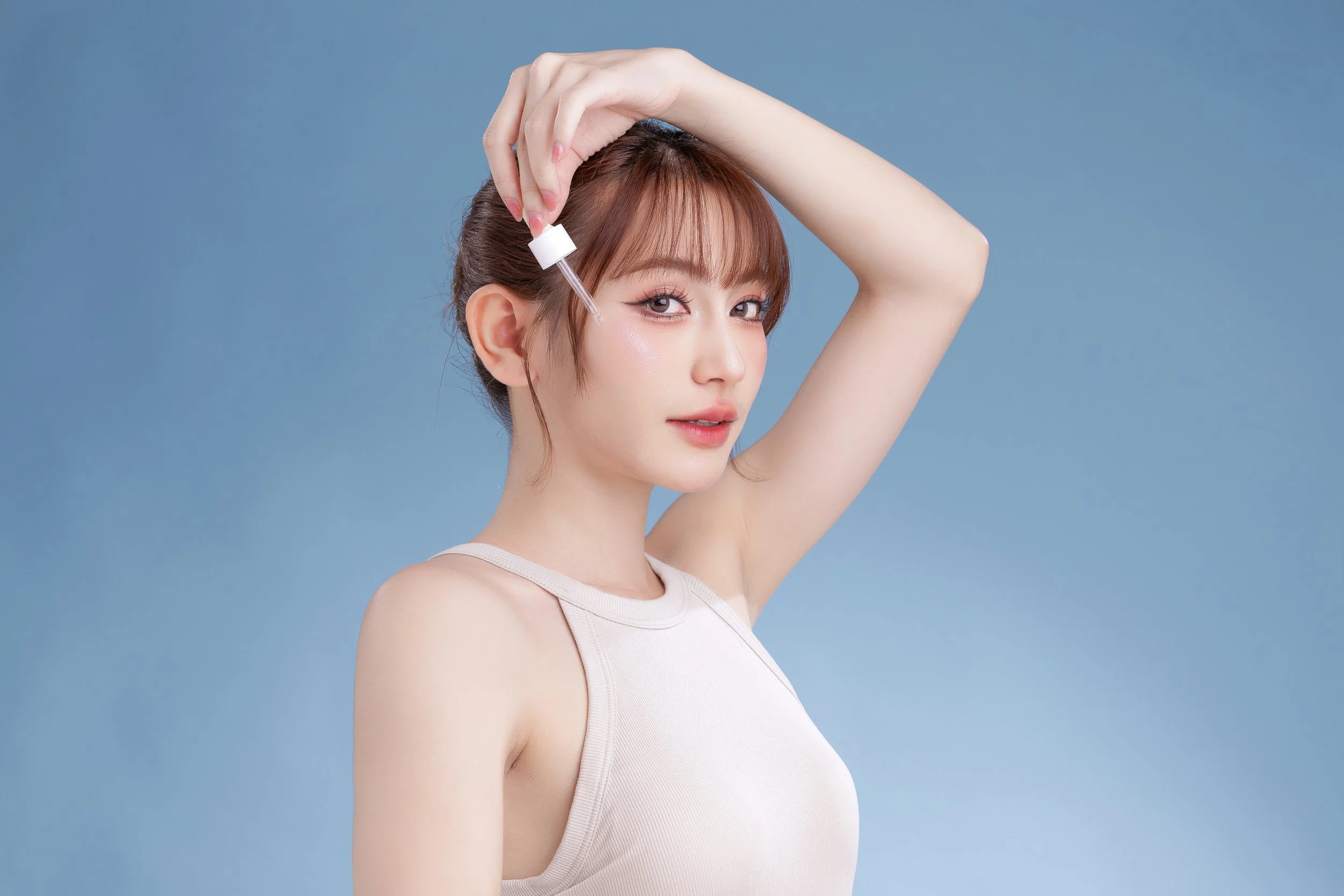 A young woman applying skincare serum to her face with a dropper, against a soft blue background.