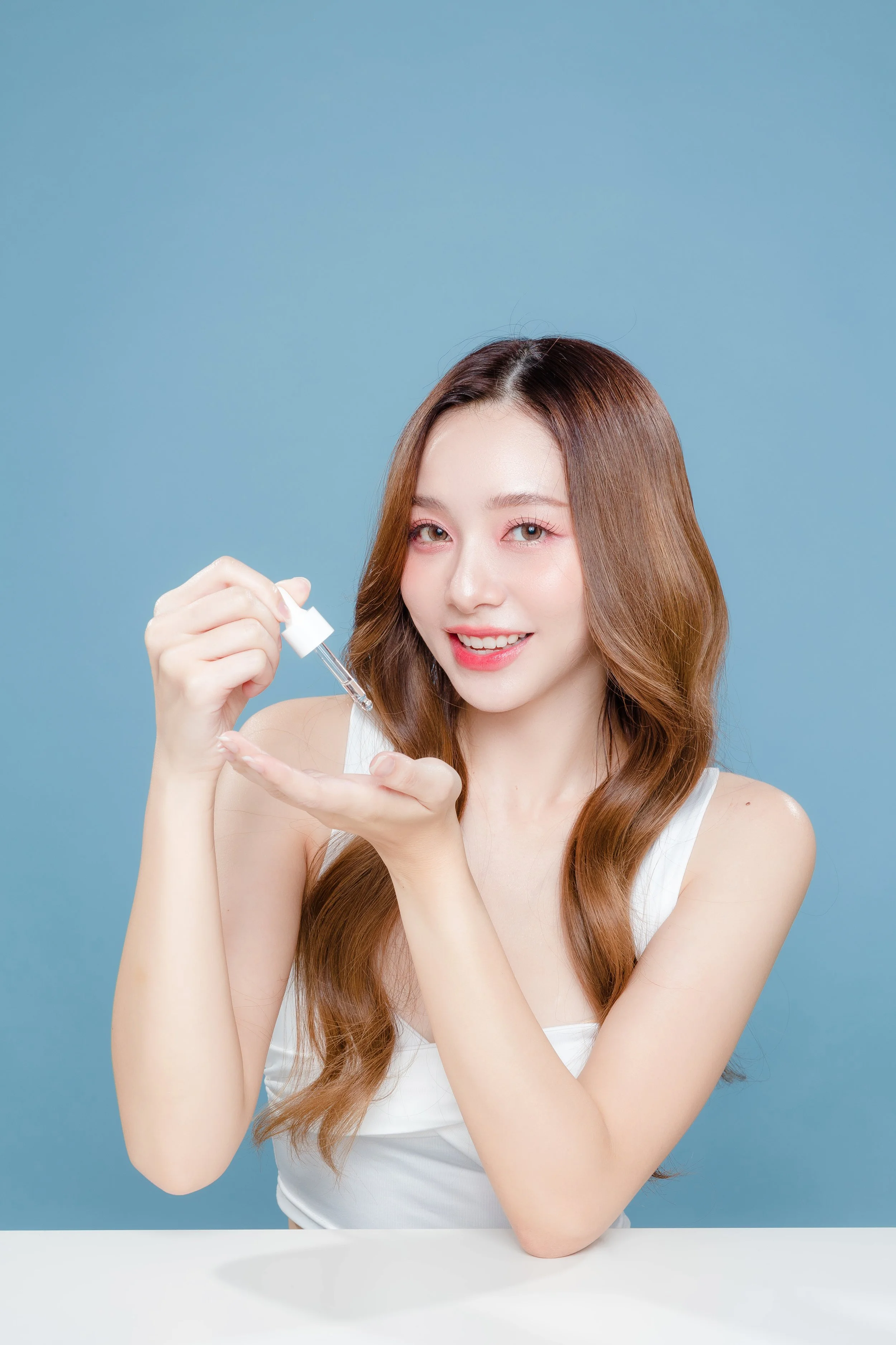 Young woman with long wavy hair and a white top holding a dropper and applying skincare to her hand against a blue background.